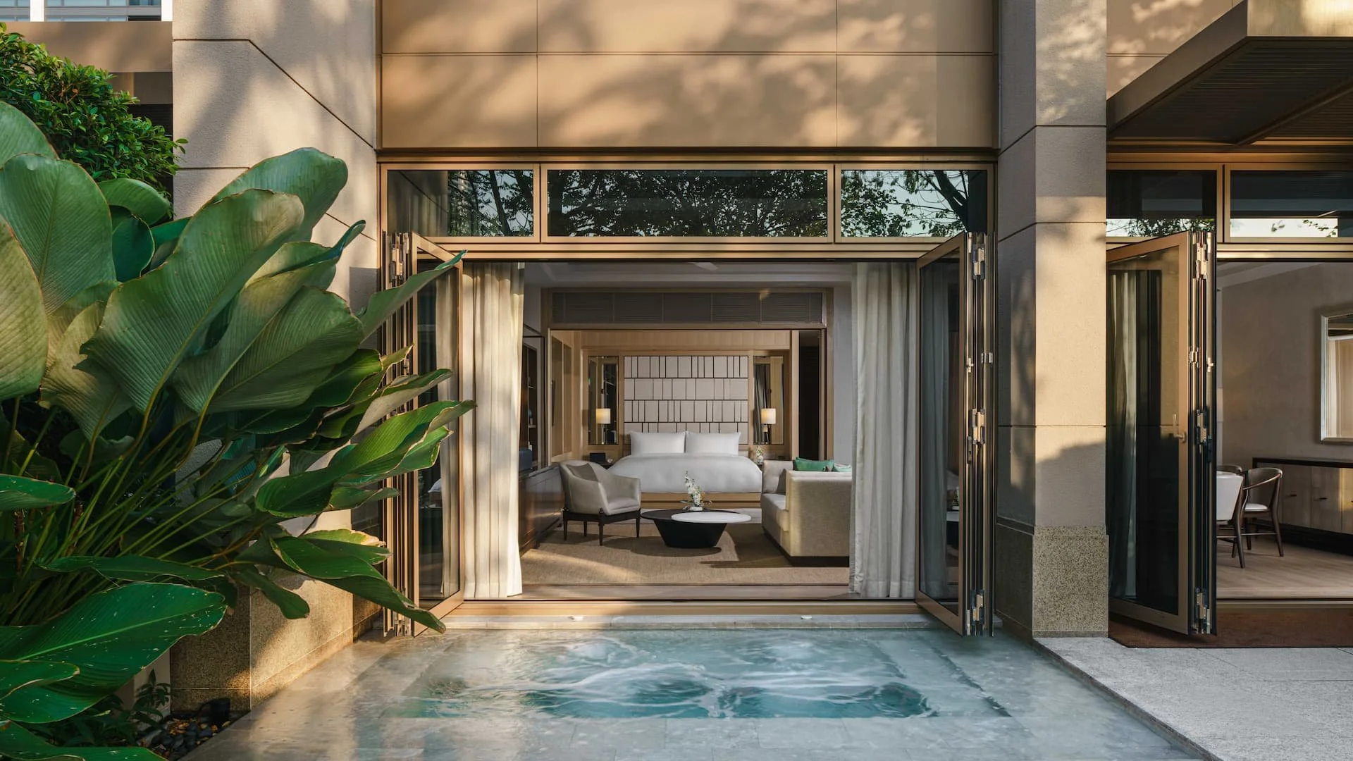 View of a luxury hotel room with a Jacuzzi outside, leading into a bedroom with a large bed and sitting area, surrounded by large green plants and soft lighting.