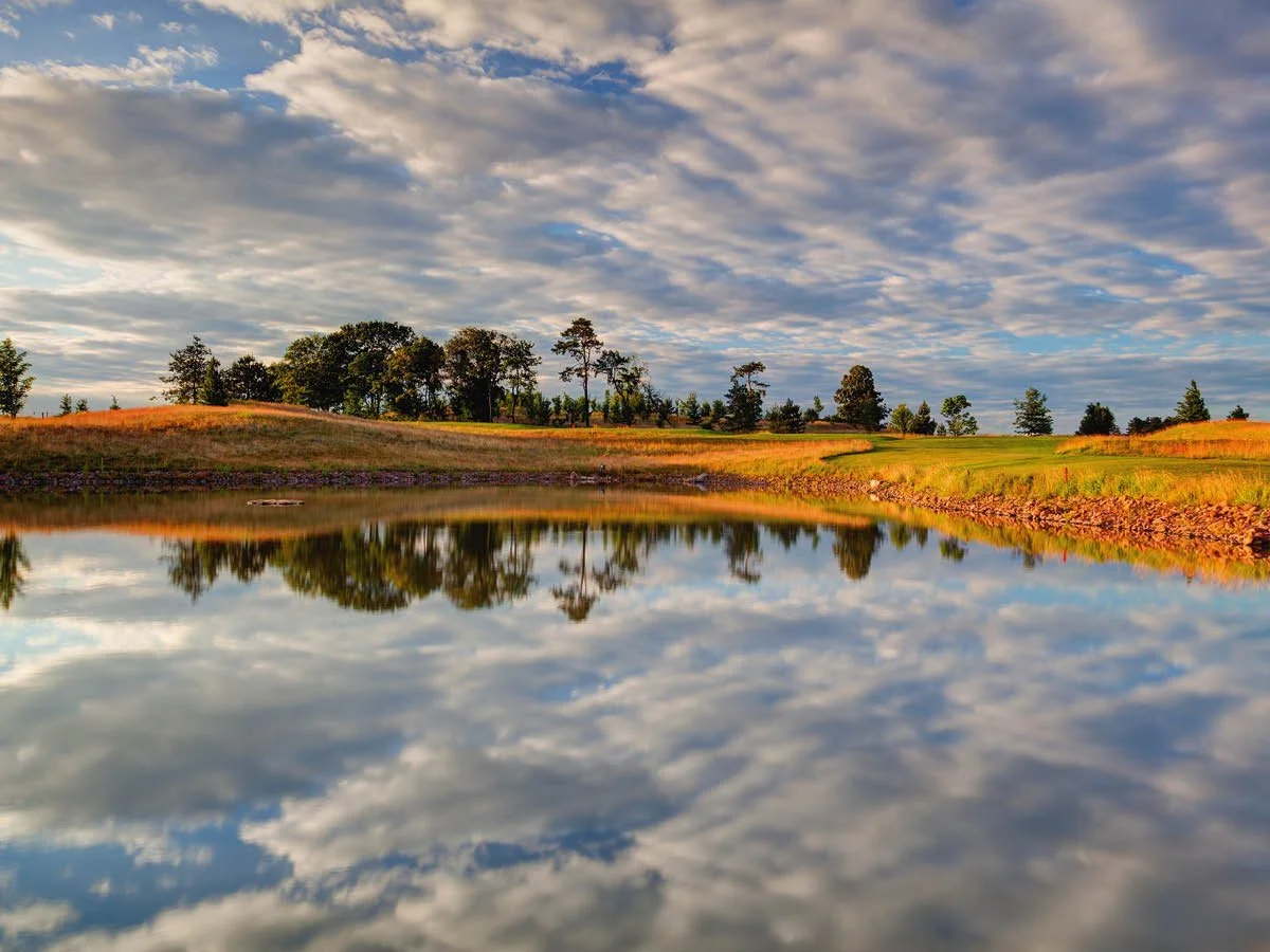 A scenic landscape of a calm lake reflecting a partly cloudy sky and distant trees with grassy hills on either side.