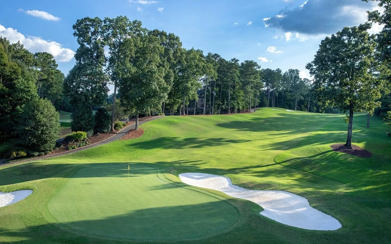 A golf course with a putting green, sand bunkers, and a flagstick, bordered by trees under a partly cloudy sky.