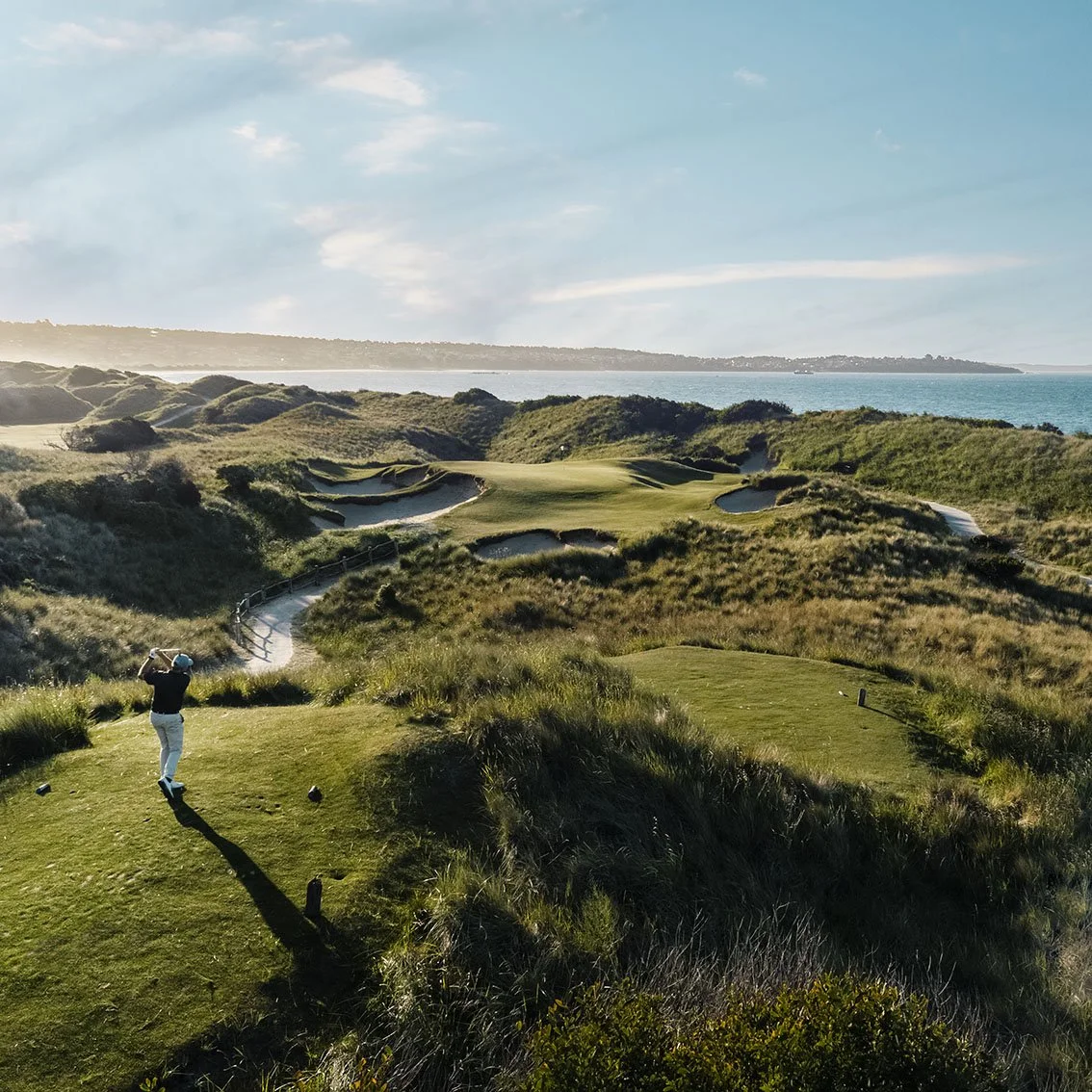A golfer playing on a scenic coastal golf course with sand traps and greenery, overlooking the ocean under a partly cloudy sky.