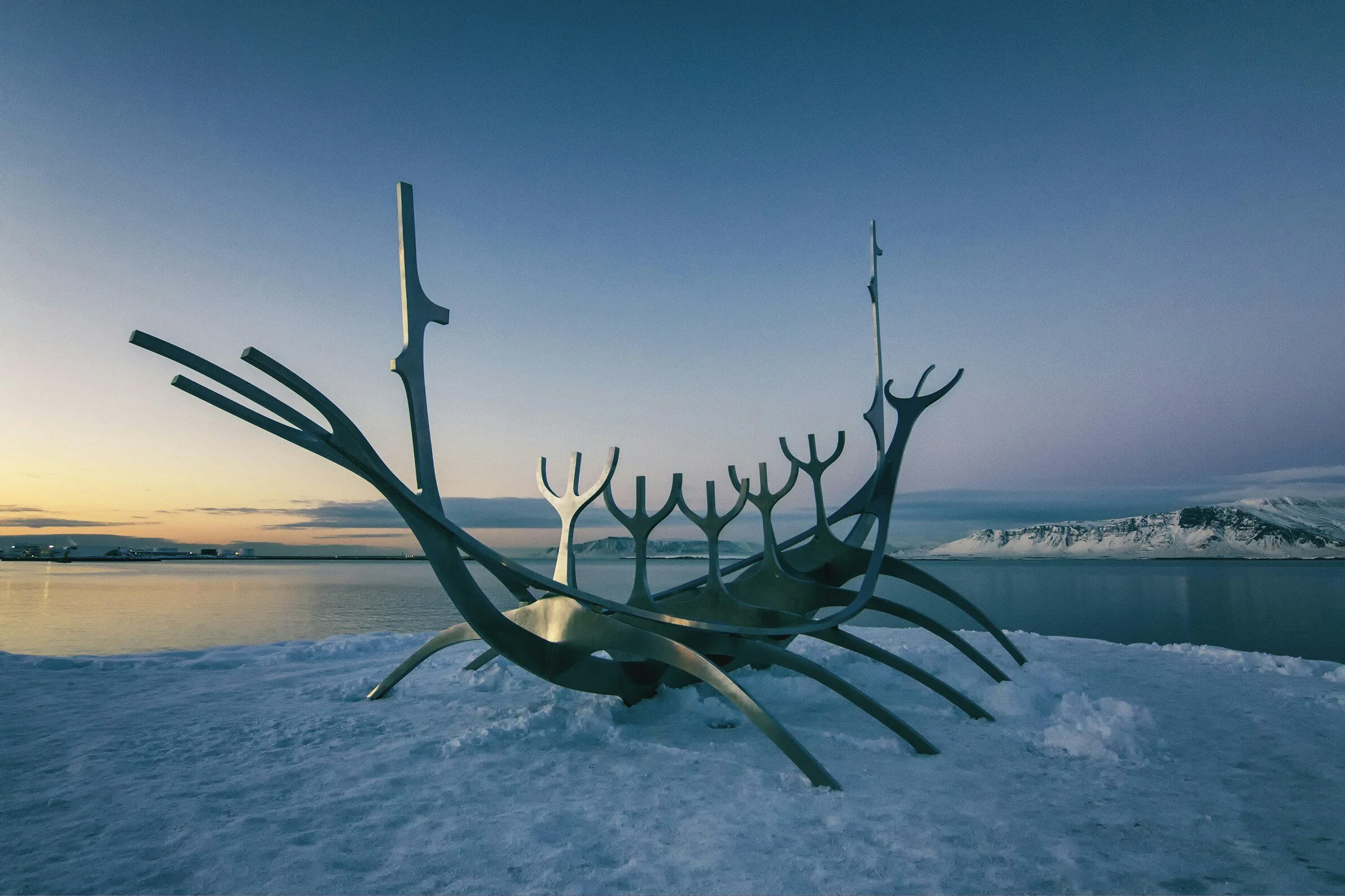 Large metallic sculpture resembling a whale's skeleton in a snowy landscape with a body of water and snow-covered mountains in the background, under a twilight sky.