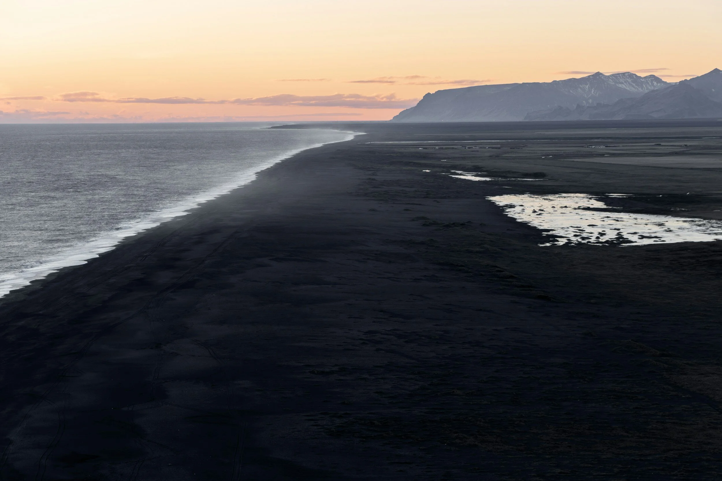 A coastal landscape with a dark volcanic sand beach, shallow pools, and a mountain range in the distance at sunset.