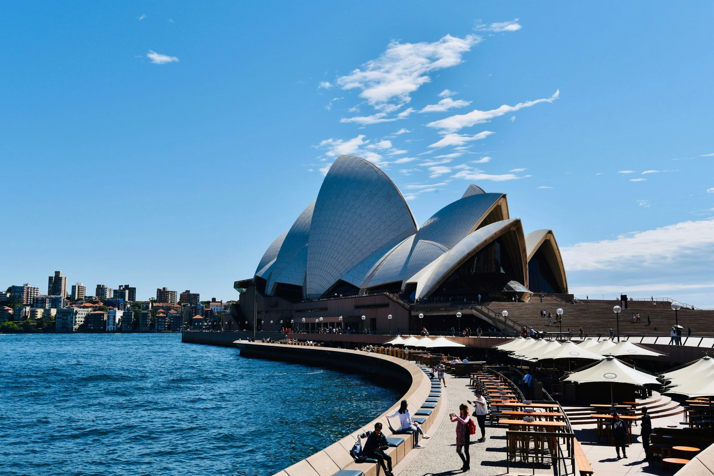 Sydney Opera House with people sitting and walking along the waterfront, under a blue sky with clouds.