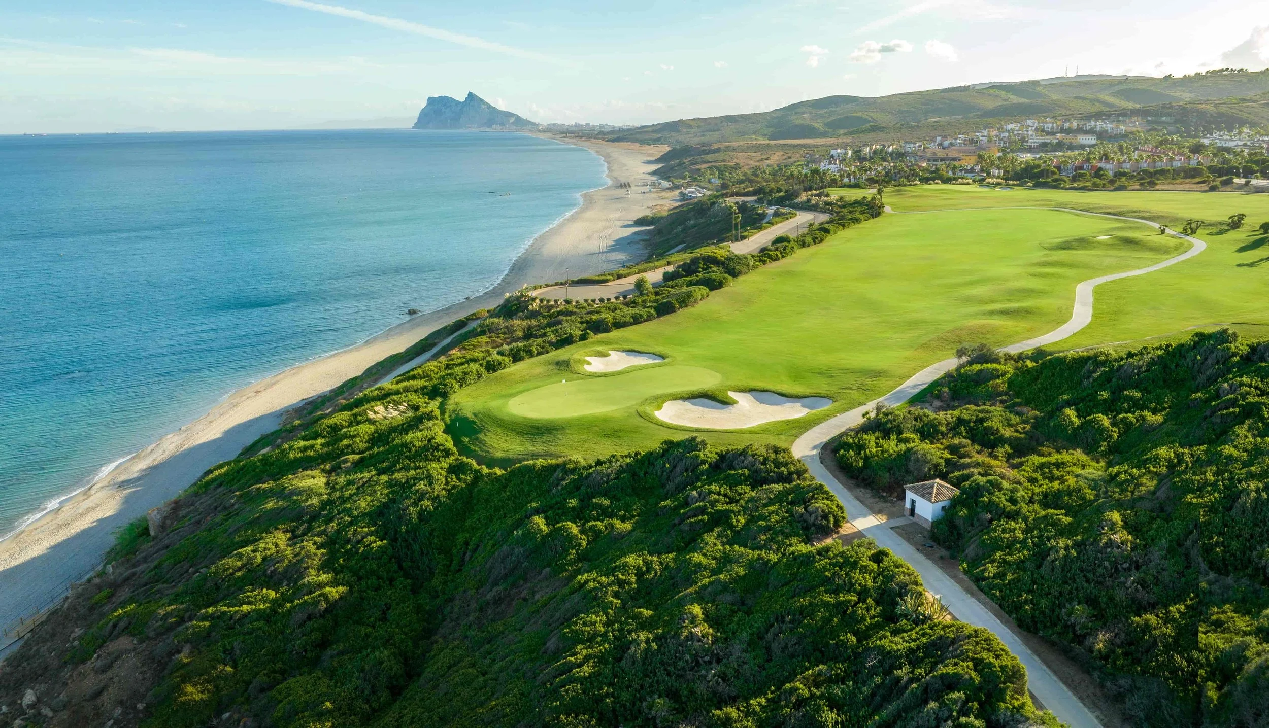 Golf course with sand traps, a house, trees, and a mountain in the background under a cloudy sky.