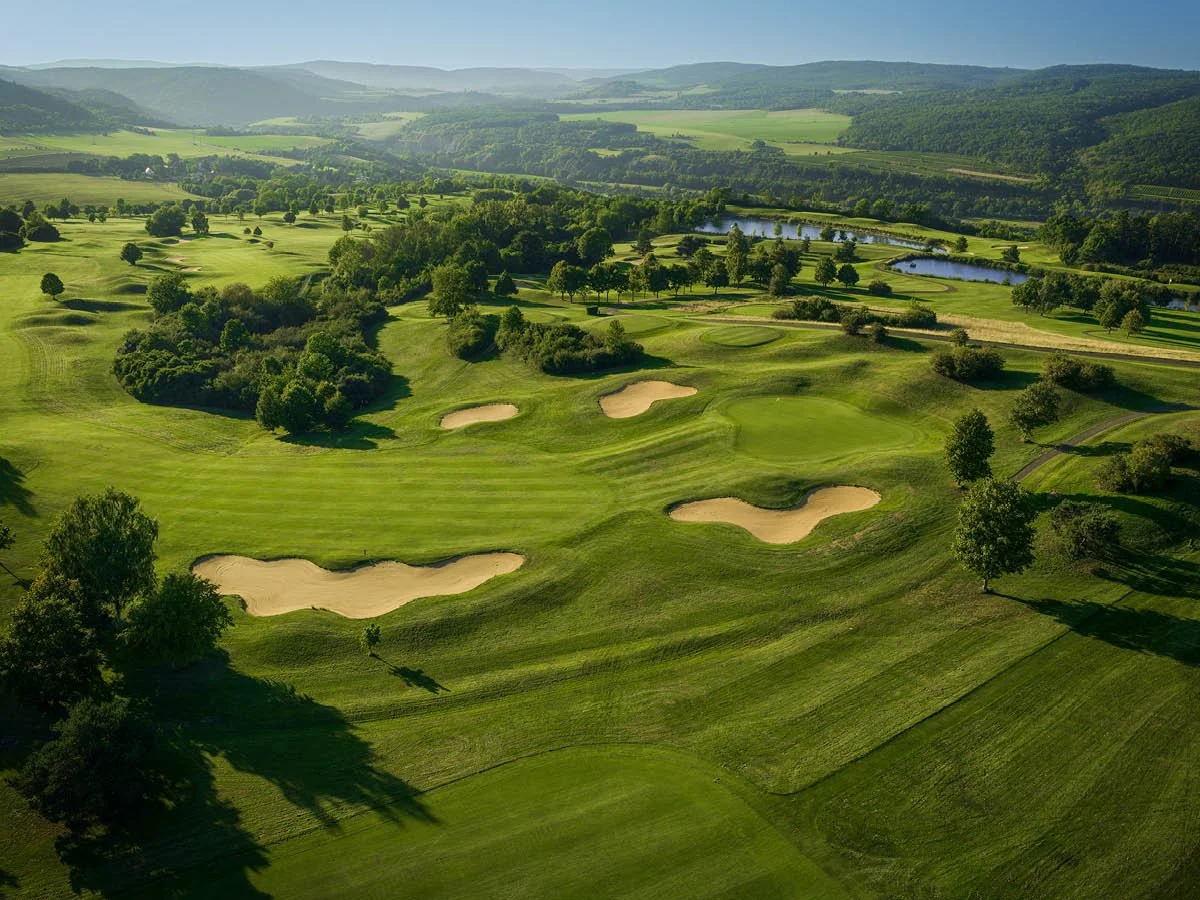 Aerial view of a lush green golf course with sand traps, trees, water features, and rolling hills in the background.