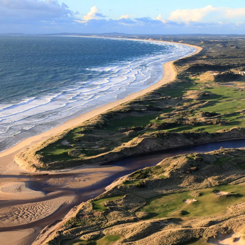 Aerial view of a golf course located along a coastline with sandy beaches and ocean waves in the background.