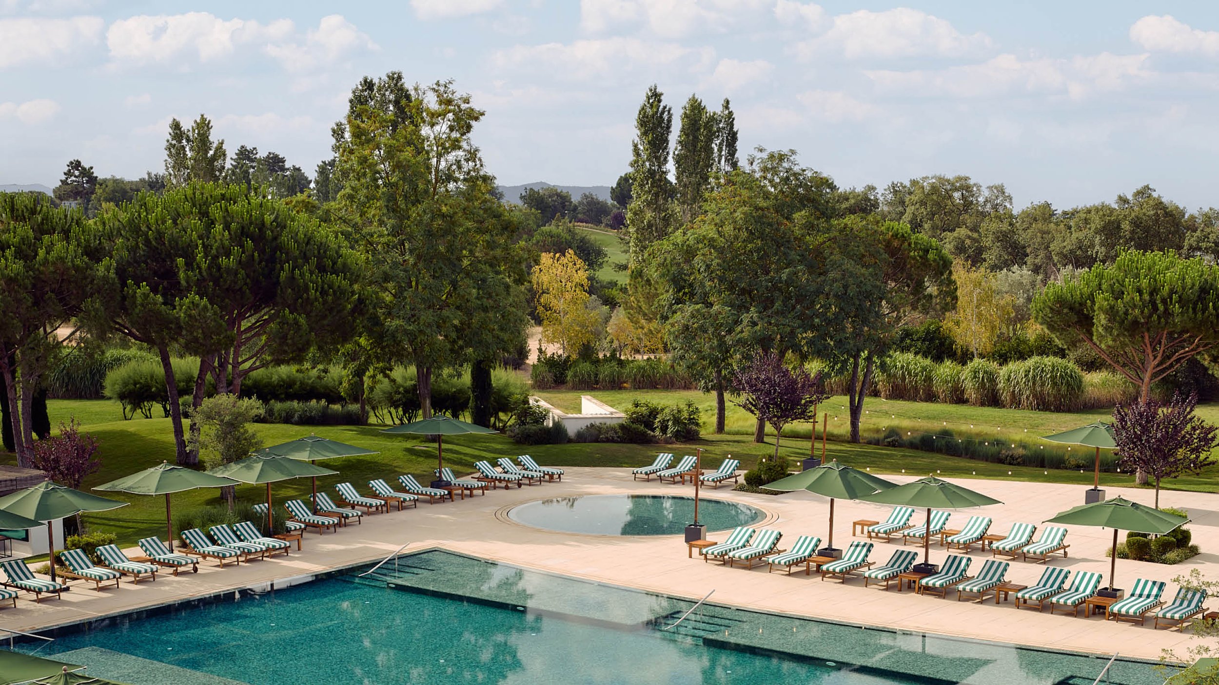 Swimming pool area with lounge chairs and green umbrellas, surrounded by trees and greenery under a partly cloudy sky.