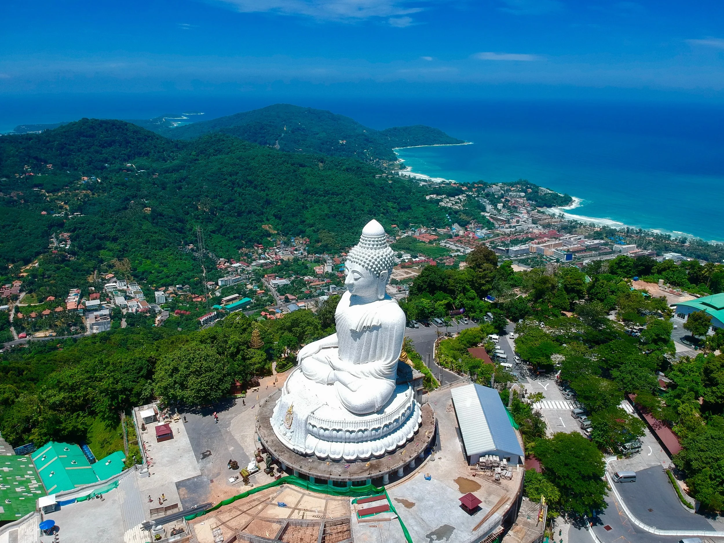 A large white Buddha statue in a seated meditation pose on a hilltop, overlooking a coastal town surrounded by lush green mountains and the ocean in the background.