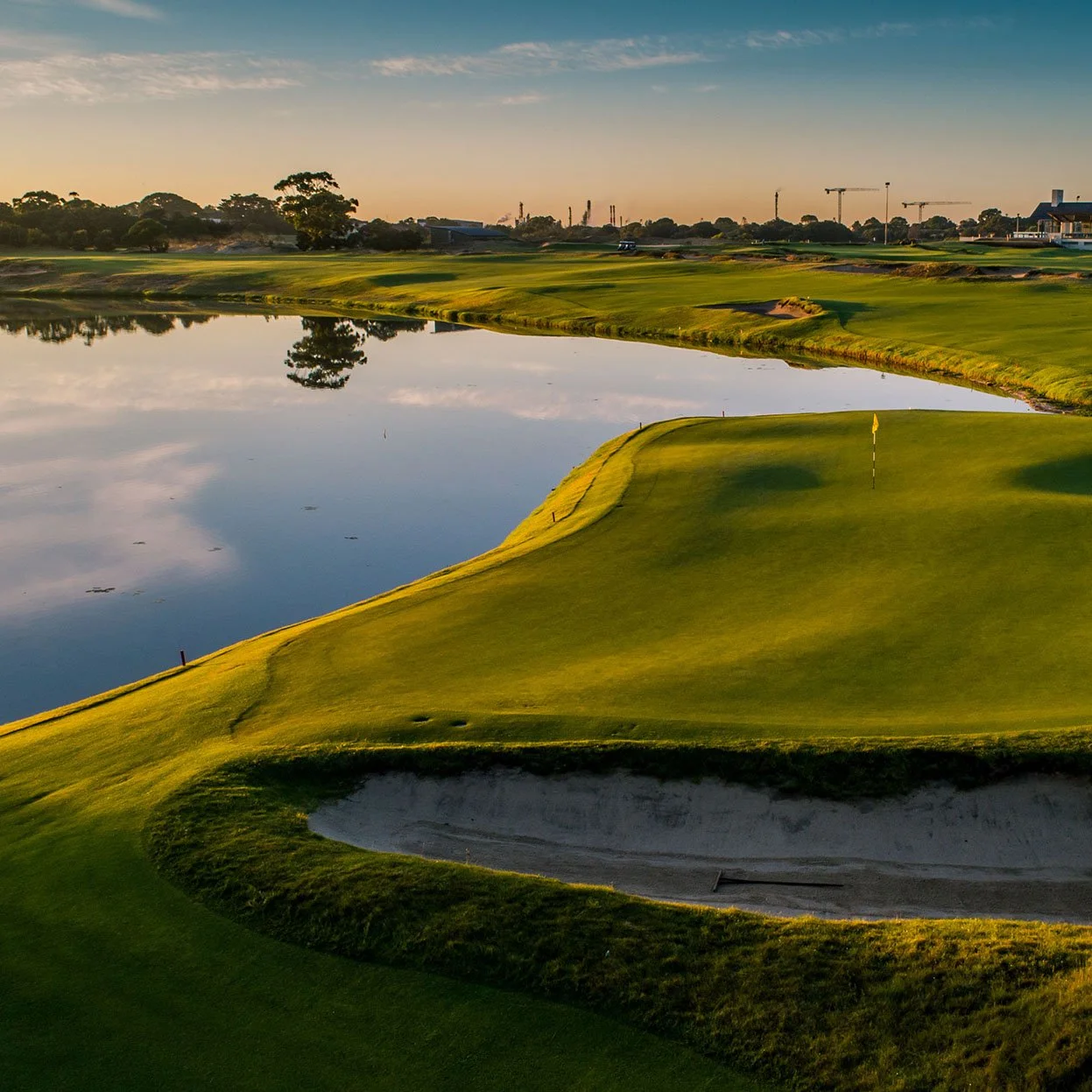 A golf course featuring a green, sand bunker, water hazard, and a flagstick under a sunset sky with construction cranes and trees in the background.