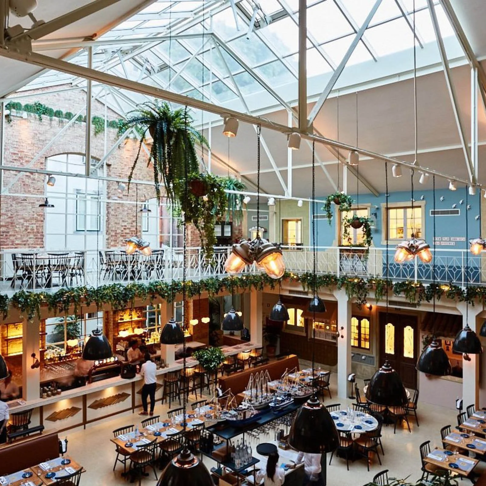 Interior of a restaurant with a high glass ceiling, hanging lights, patterned tables, and greenery decorations.