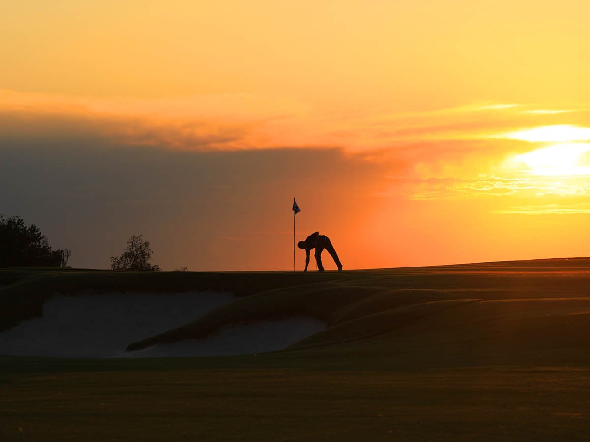 Silhouette of a person on a golf course near a flag at sunset, with a colorful sky.