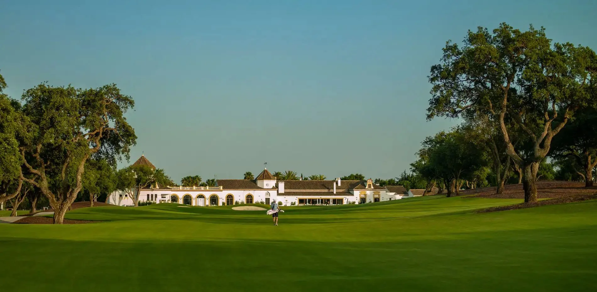A person walking on a golf course near a large white clubhouse with arches, towers, and a tiled roof, surrounded by trees and greenery.