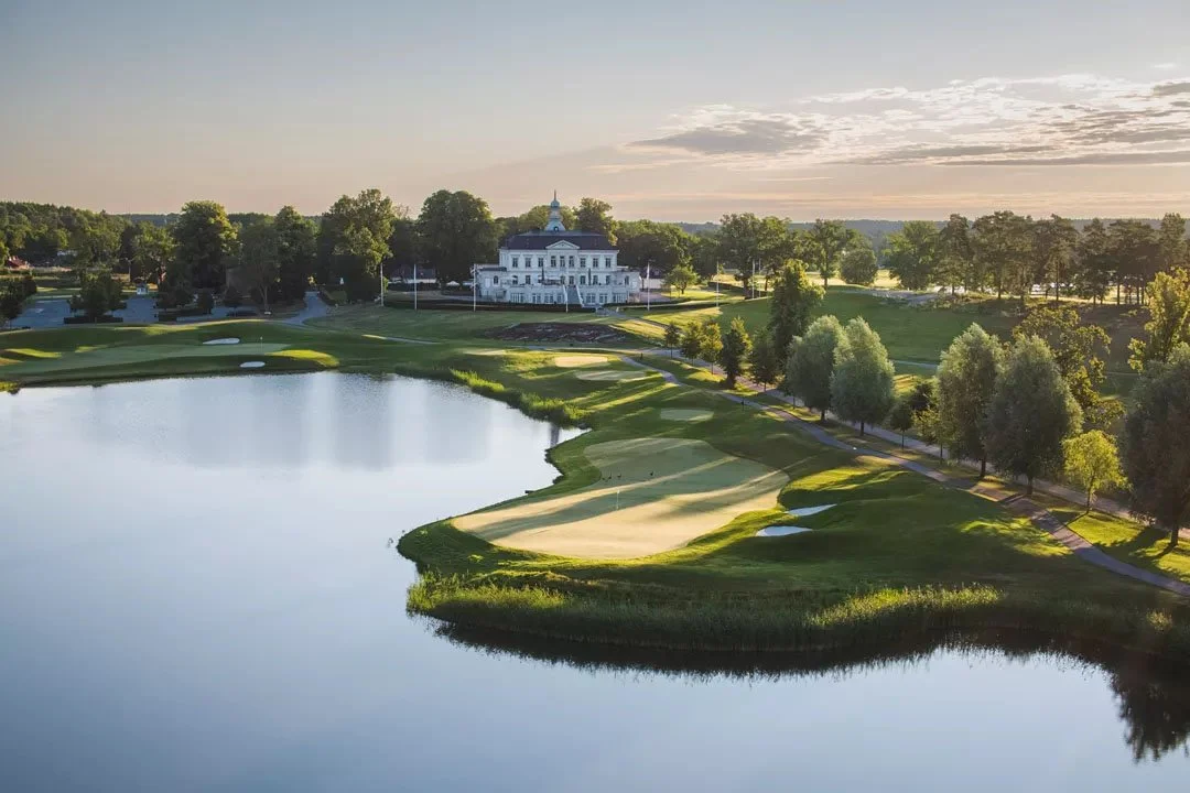 A scenic view of a golf course with a water hazard, sand traps, green fairways, and a clubhouse in the background during sunset.