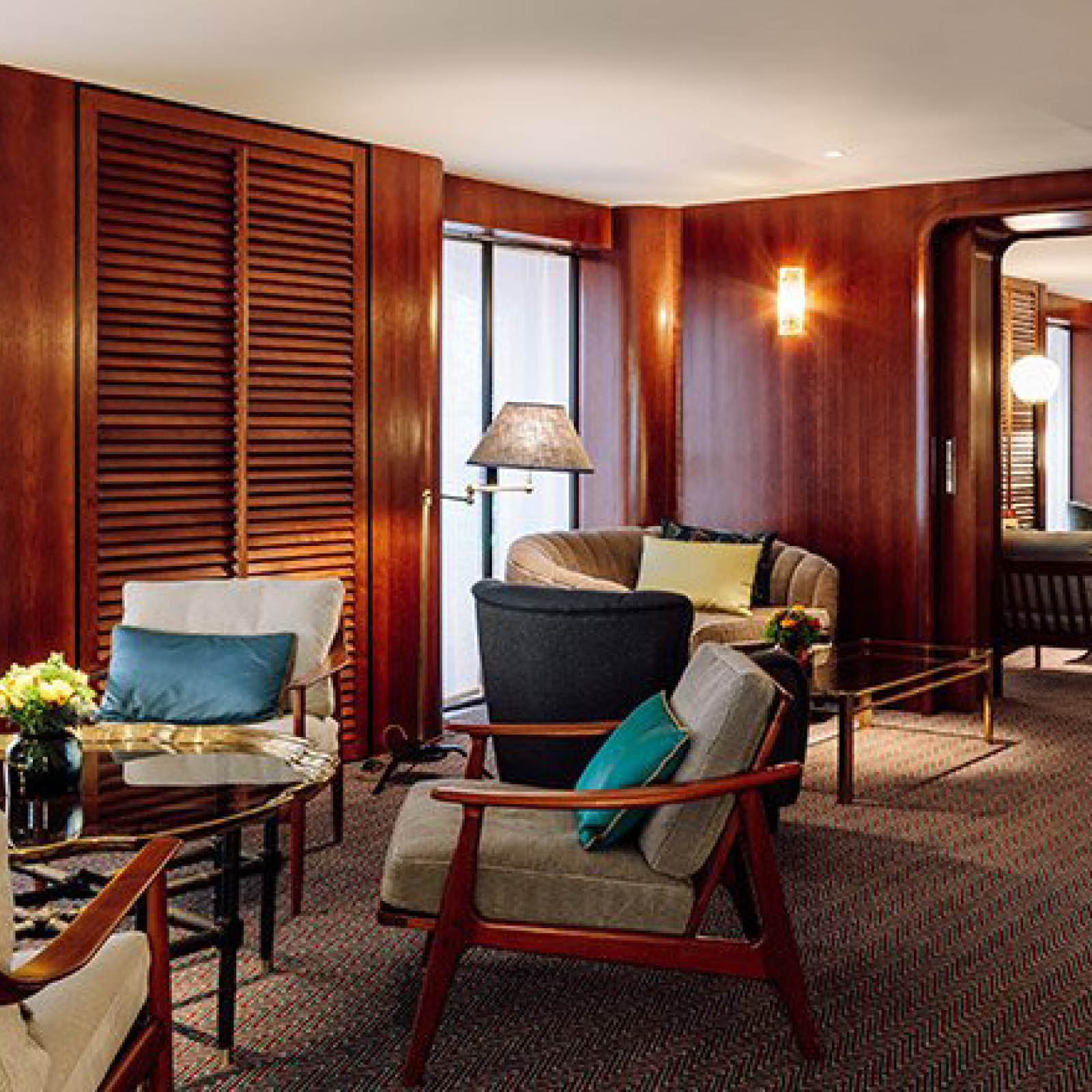 Elegant hotel living room with wooden wall paneling, comfortable chairs with cushions, a glass coffee table with a flower arrangement, and large windows providing natural light.