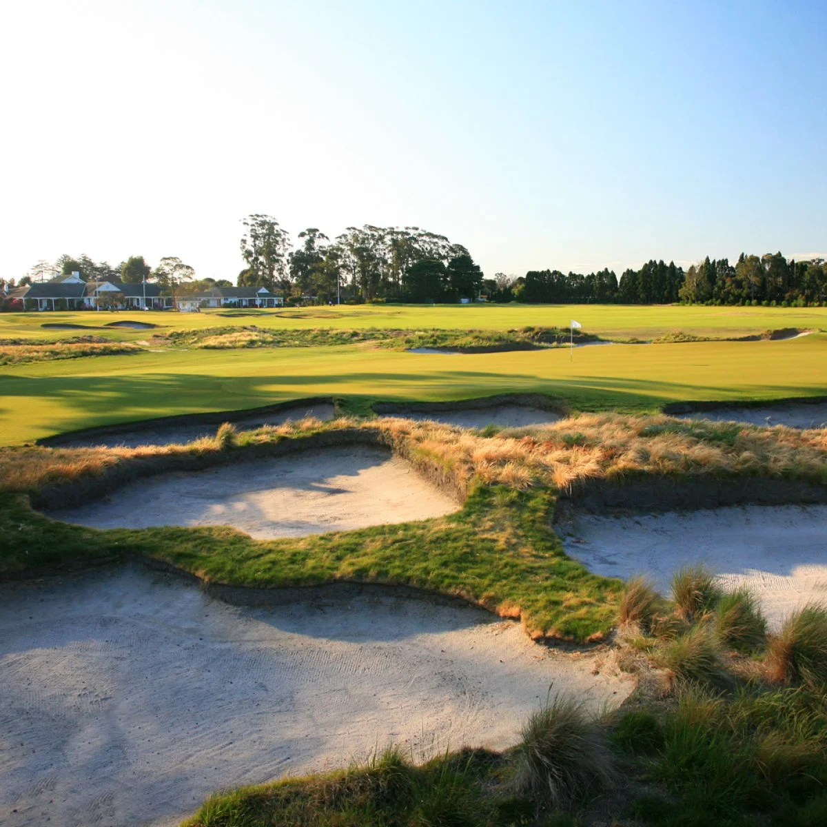 Aerial view of a golf course with well-maintained green, sand bunkers, trees, and distant city skyline at sunset.