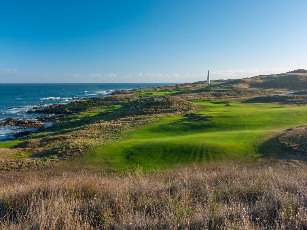 Coastal golf course with grassy fairways and dunes overlooking the ocean, with a lighthouse in the distance under a clear blue sky.