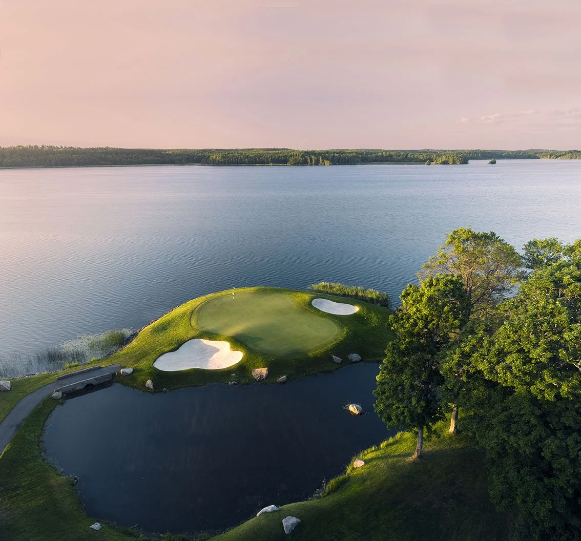 Aerial view of a golf course with a green, sand traps, water hazards, trees, and a large lake in the background during sunset.