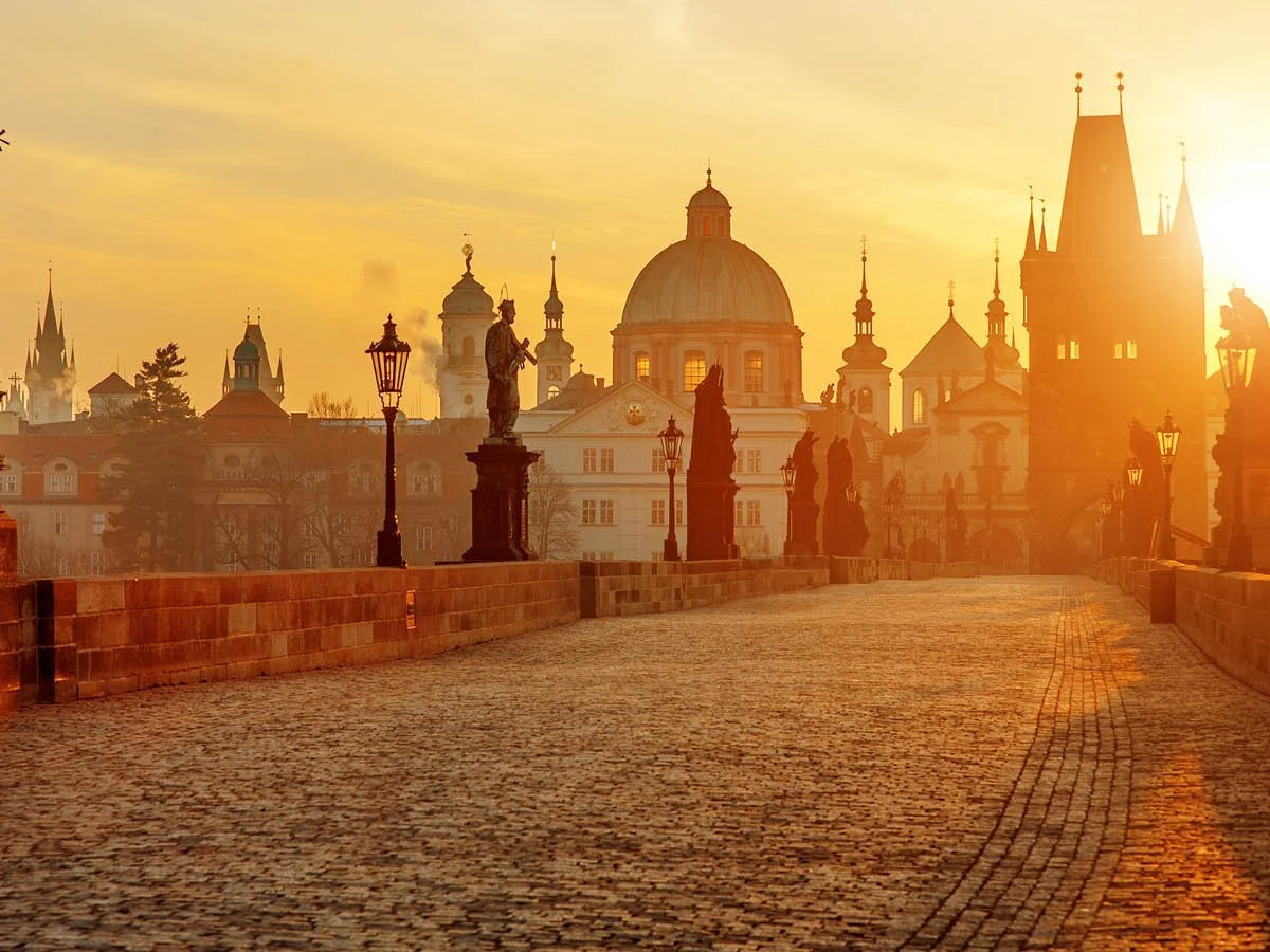 Sunset view over Charles Bridge in Prague with statues, historic buildings, and a large domed church in the background.