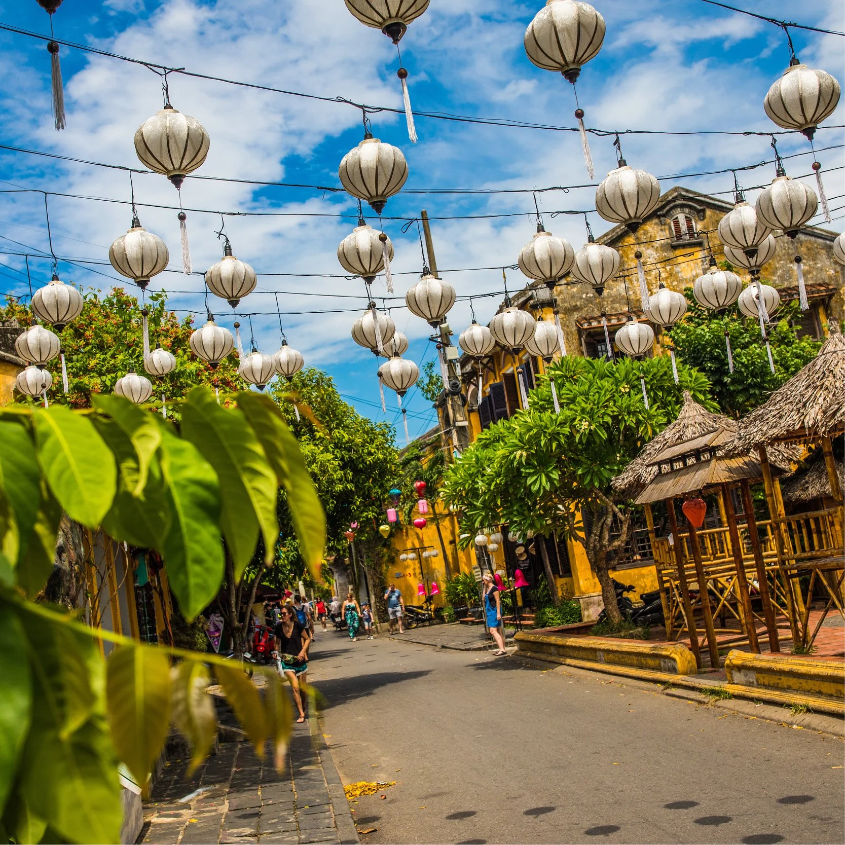 A vibrant street scene decorated with hanging paper lanterns, with people walking and buildings with rustic architecture, lush greenery, and a bright blue sky.