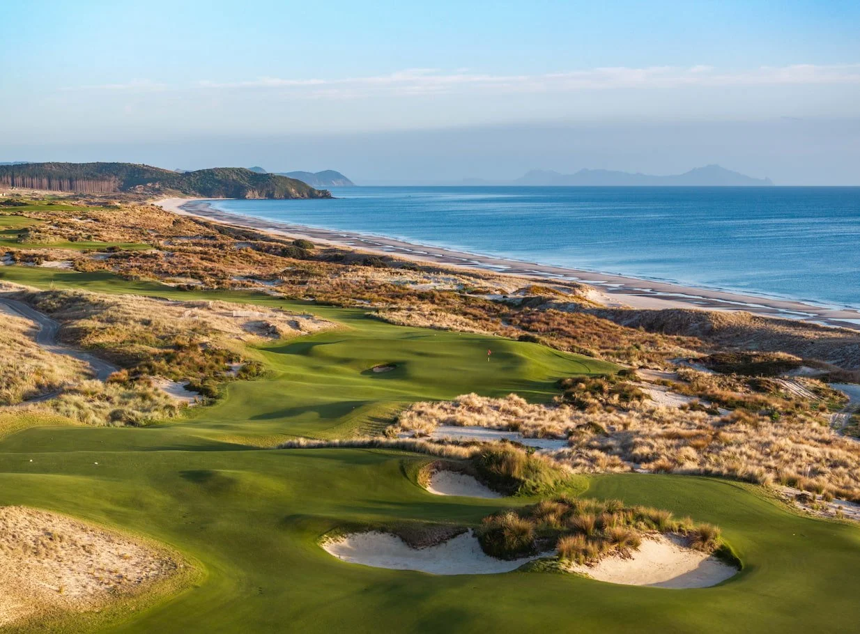 Aerial view of a sandy beach and ocean waves with a green golf course and forested hills in the background
