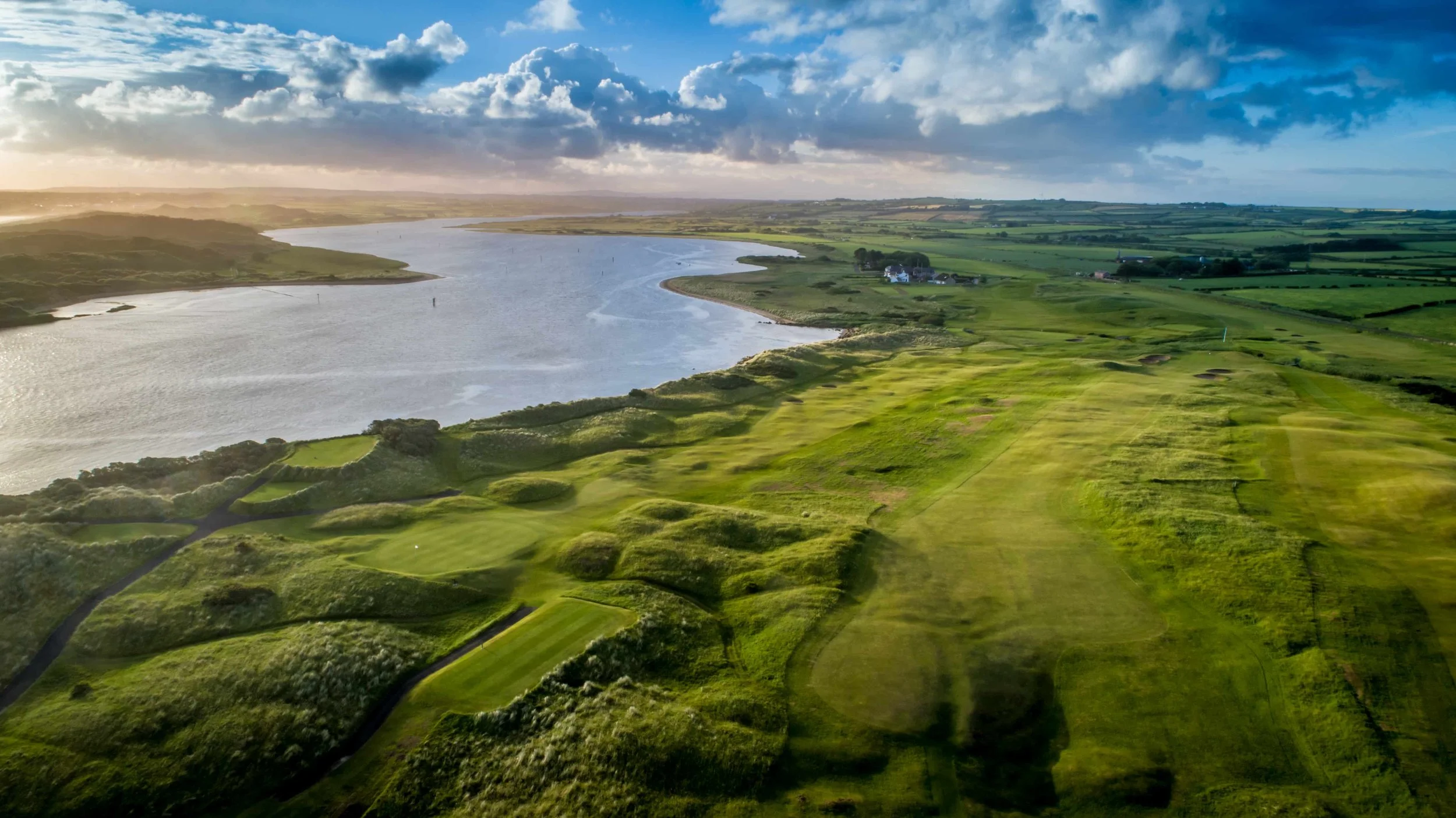 A scenic golf course on a hillside overlooking the ocean with green fairways and putting greens, under a partly cloudy sky.