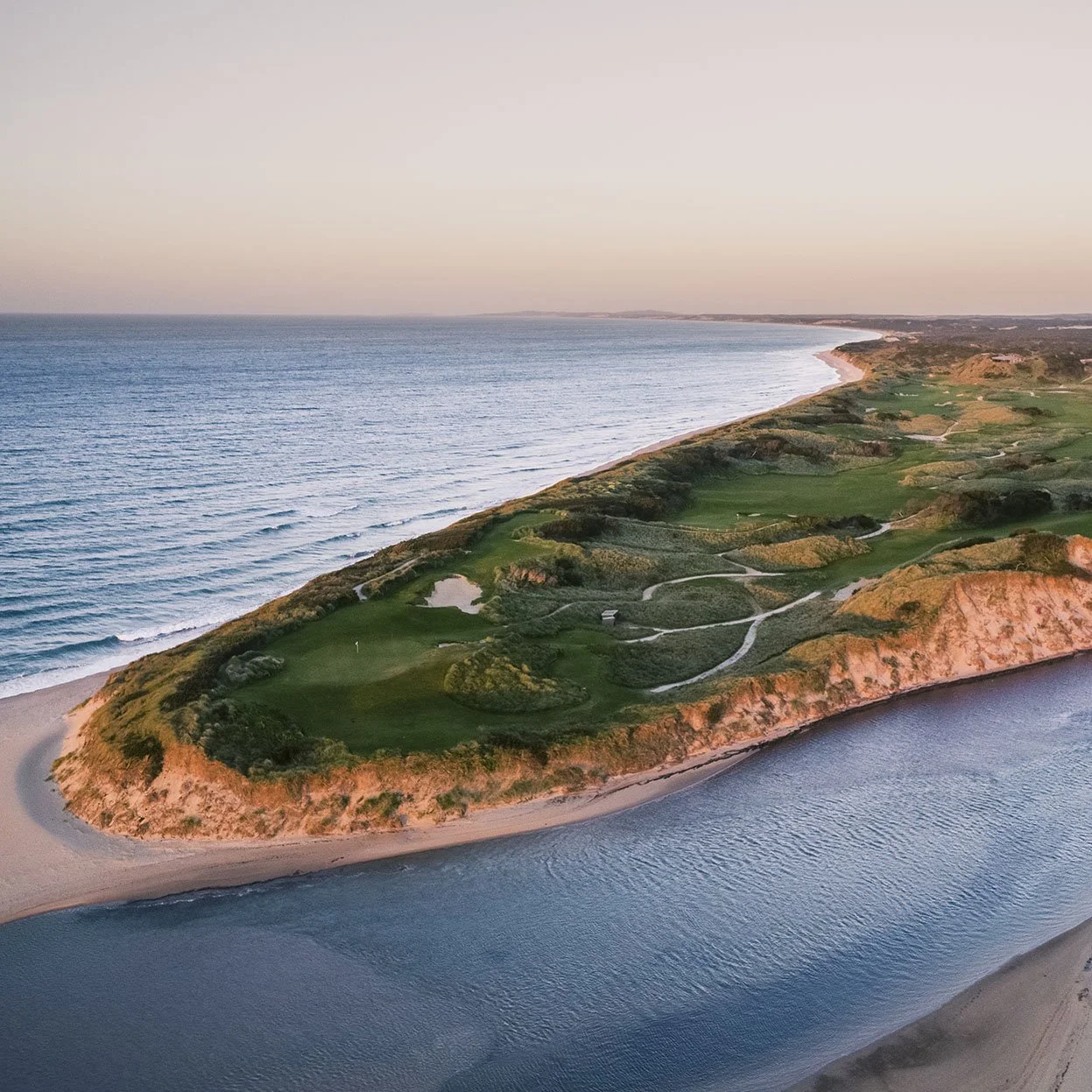 Aerial view of a coastal golf course on a narrow peninsula, with the ocean on one side and water along the other, under a clear sky at sunset.