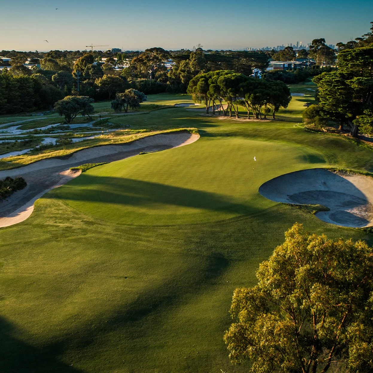 Aerial view of a golf course with well-maintained green, sand bunkers, trees, and distant city skyline at sunset.