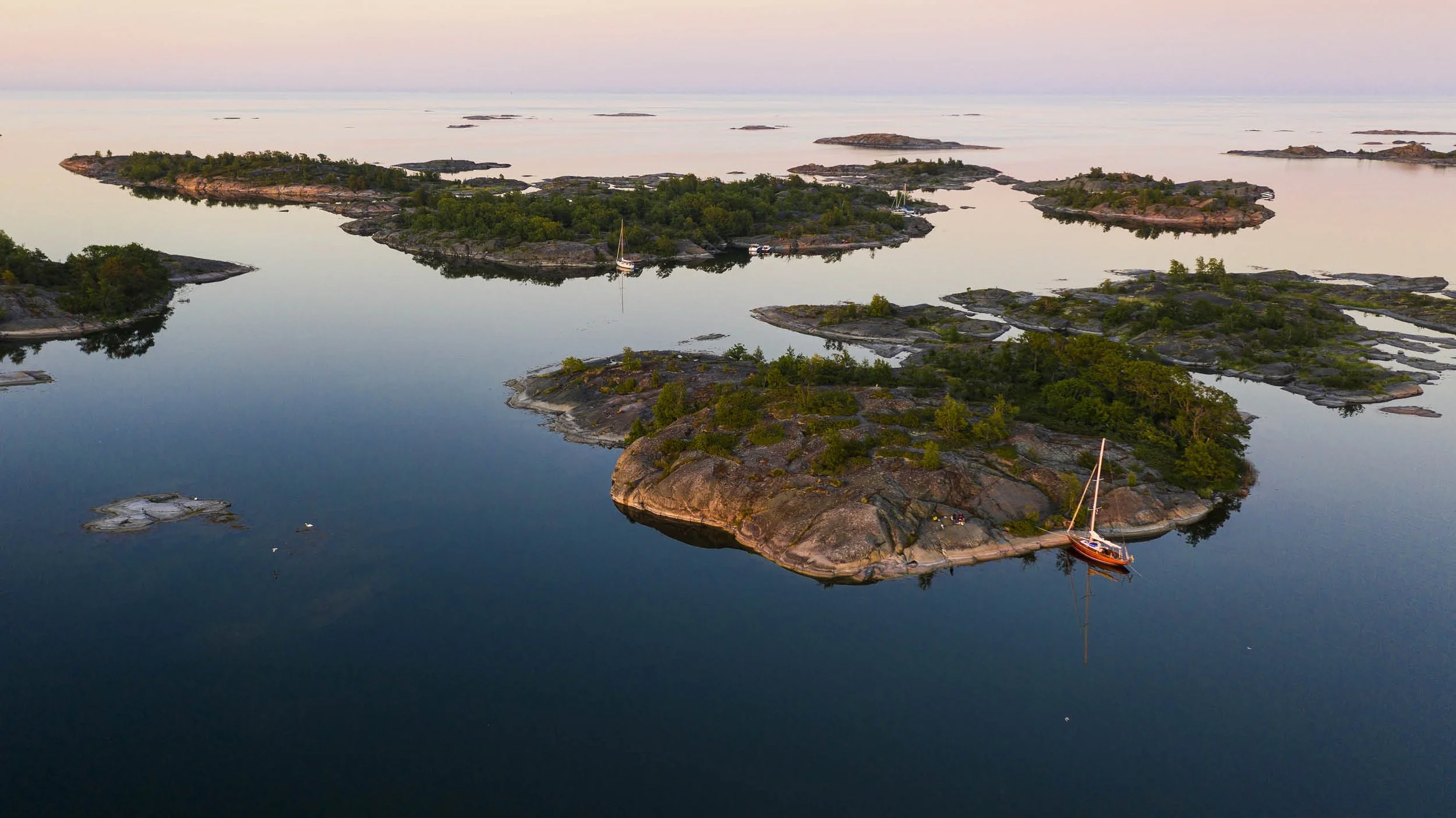 Aerial view of rocky islands with green trees in a calm body of water, with sailboats anchored near the islands, during dusk or dawn with soft lighting and pastel sky.