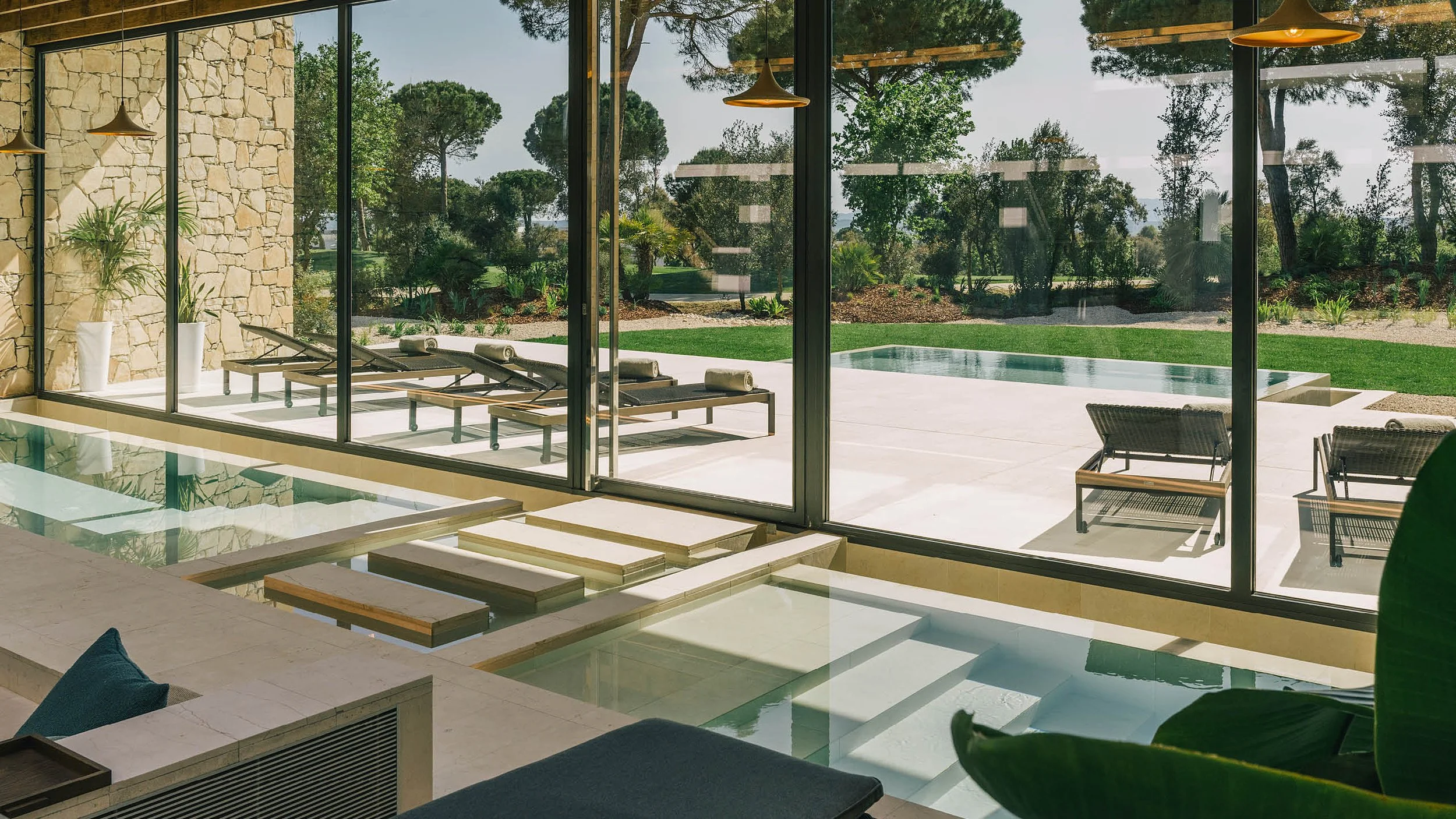 Sunny outdoor pool area with lounge chairs and lush green landscaping, viewed through large glass windows from inside a modern house.