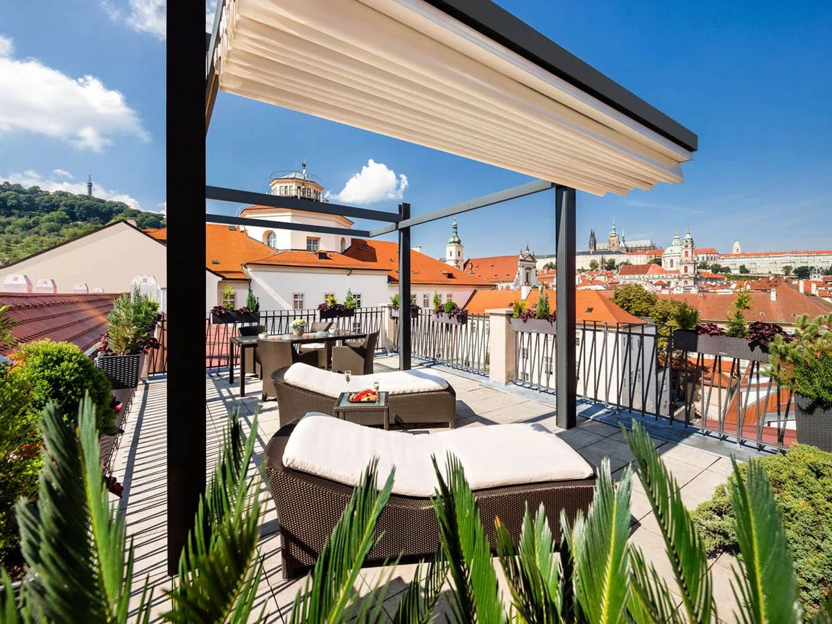 Rooftop terrace with outdoor furniture and potted plants overlooking a cityscape with historic buildings and orange rooftops under a blue sky.