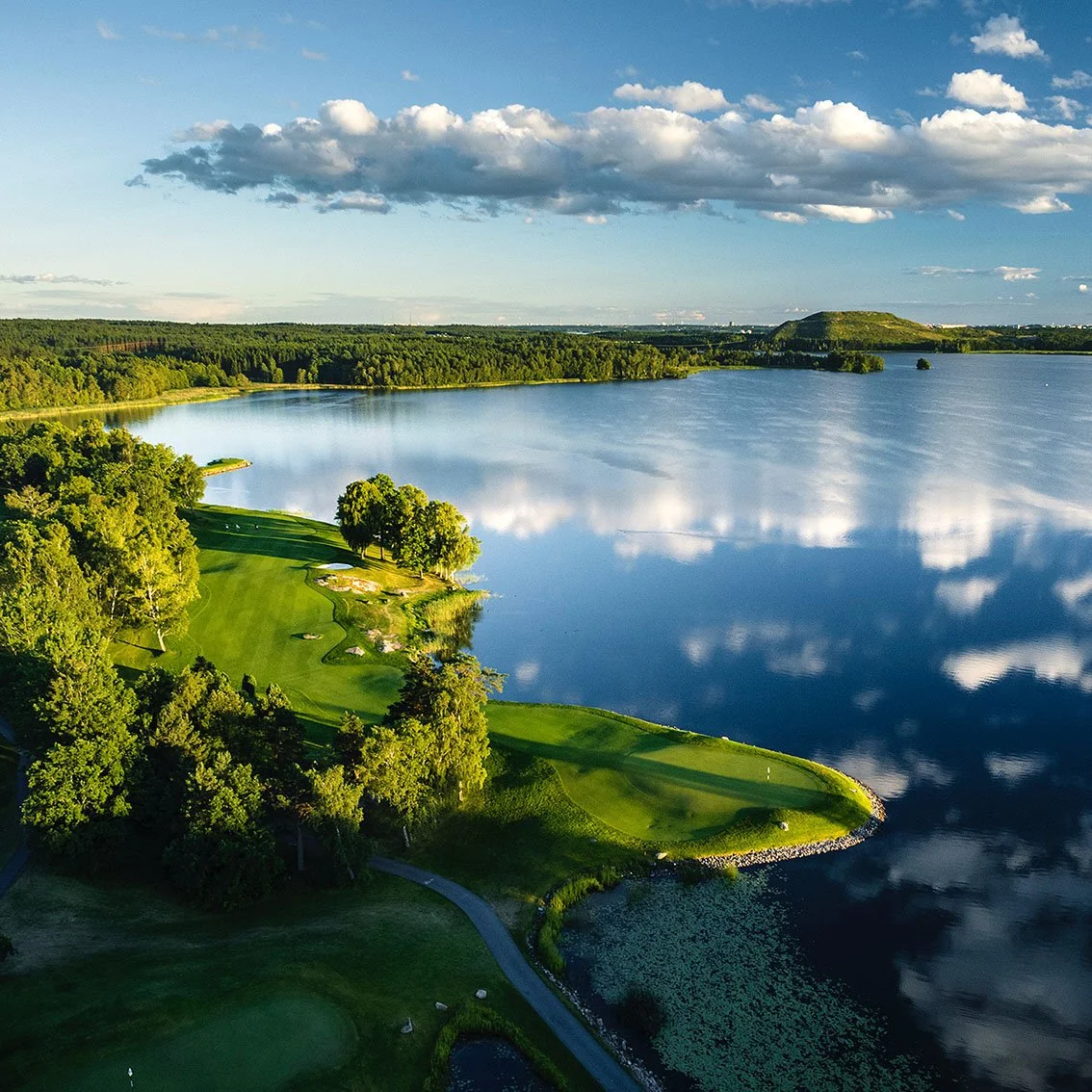 Aerial view of a golf course alongside a lake, with trees, green fairways, and a partly cloudy sky reflected in the water.