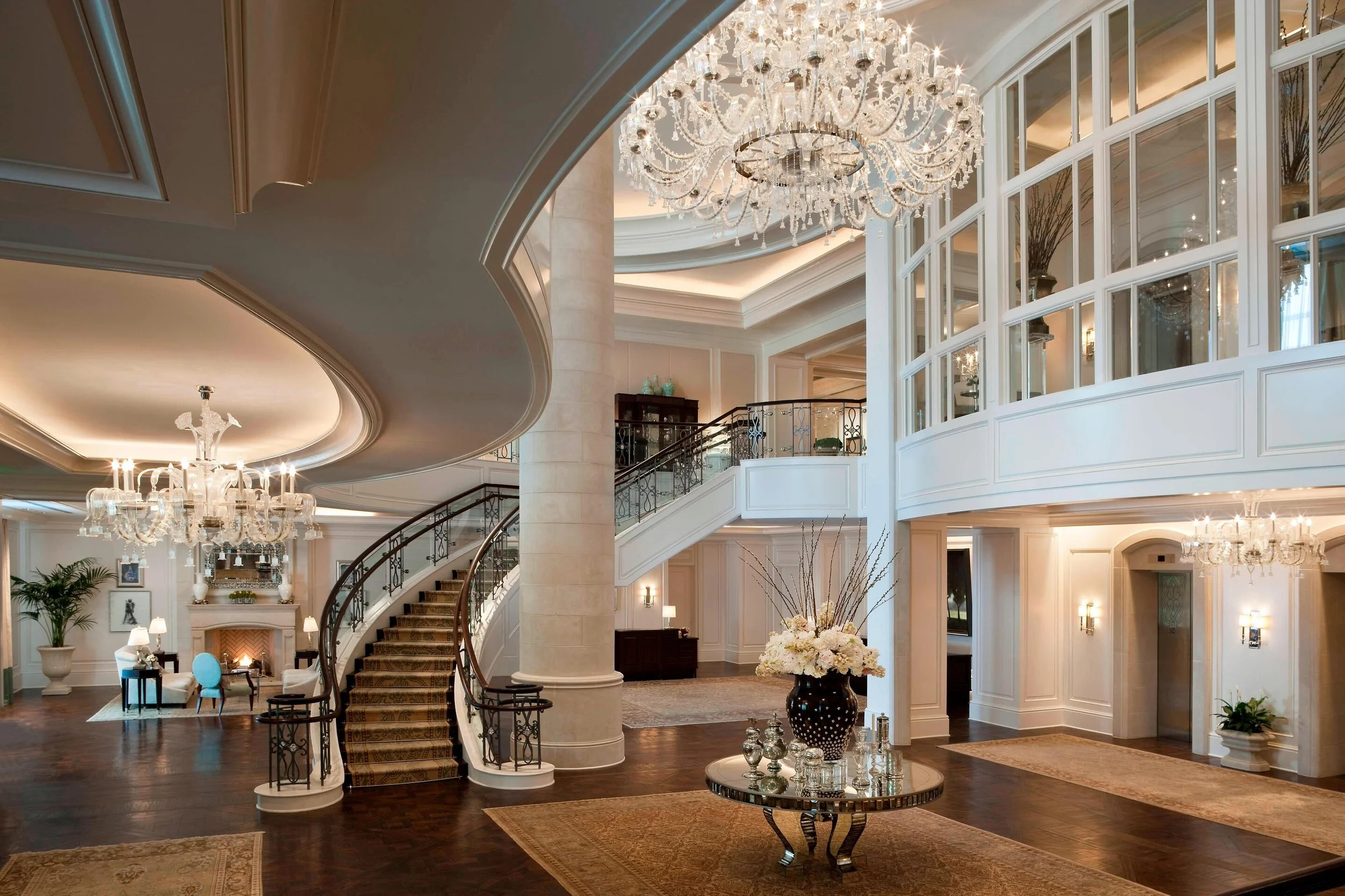 Luxurious hotel lobby with grand chandelier, white walls, dark wood flooring, elegant staircase, large floral arrangement, and cozy seating area.