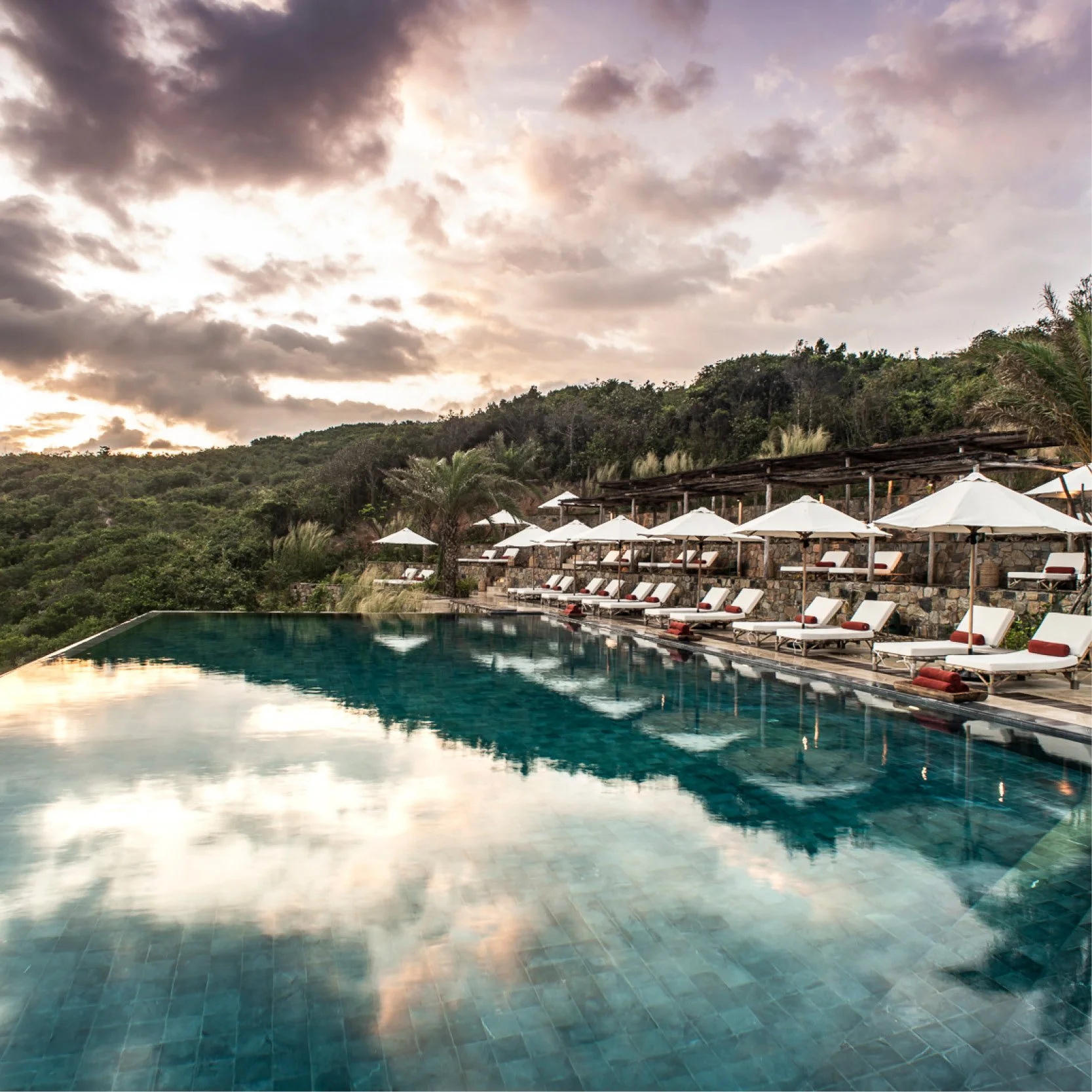 An infinity pool overlooking a lush green hillside at sunset, with white lounge chairs and umbrellas along the poolside.