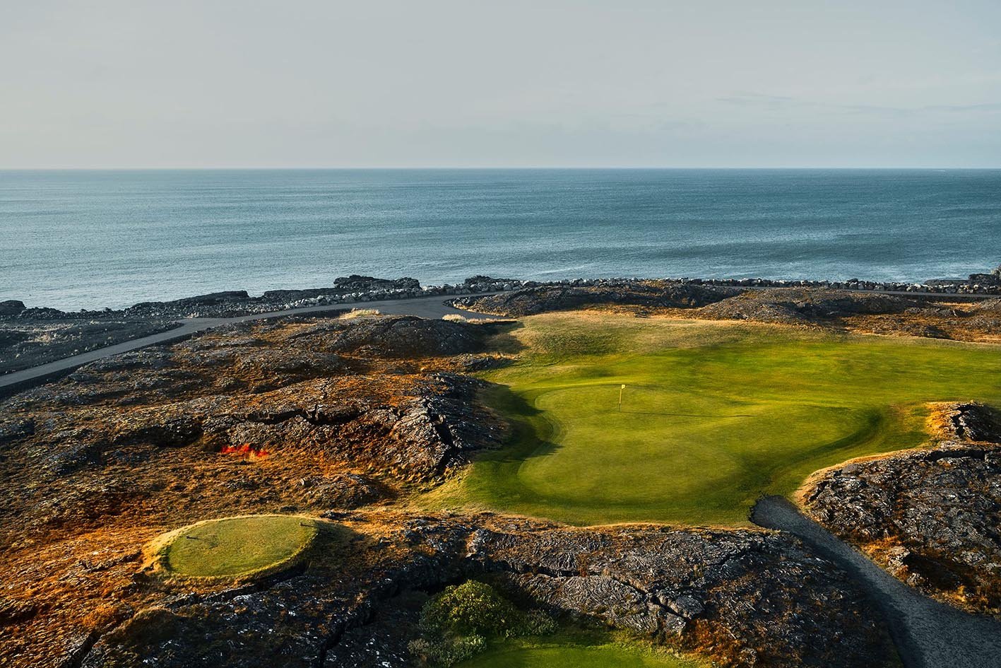 Scenic view of a green golf course surrounded by rocky terrain, with the ocean in the background under an overcast sky.