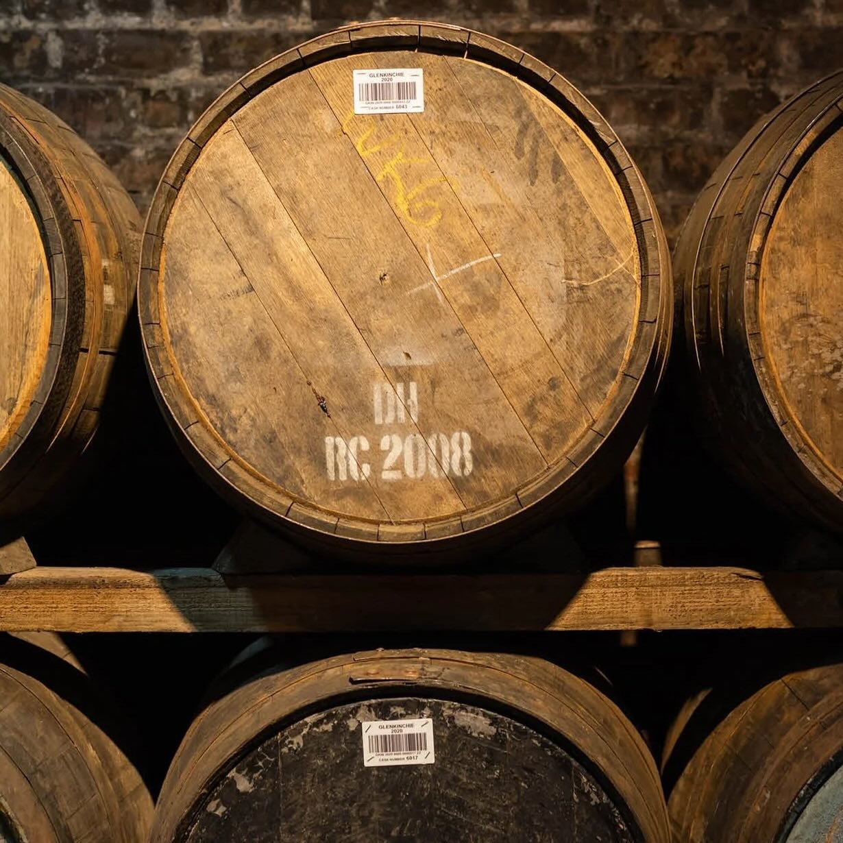 Wooden whiskey barrels stacked in storage, with one prominent barrel showing a white stencil of the label 'IN RC: 2008' and a barcode sticker on top.