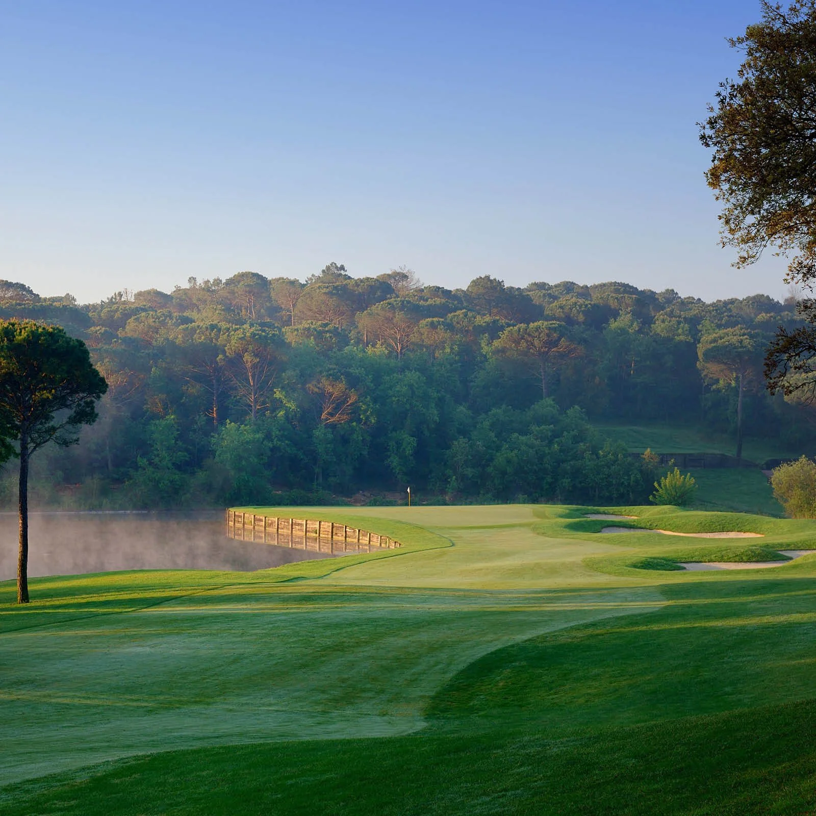 A scenic golf course with lush green grass, a small pond with mist rising from it, surrounded by trees and rolling hills in the background under a clear blue sky.