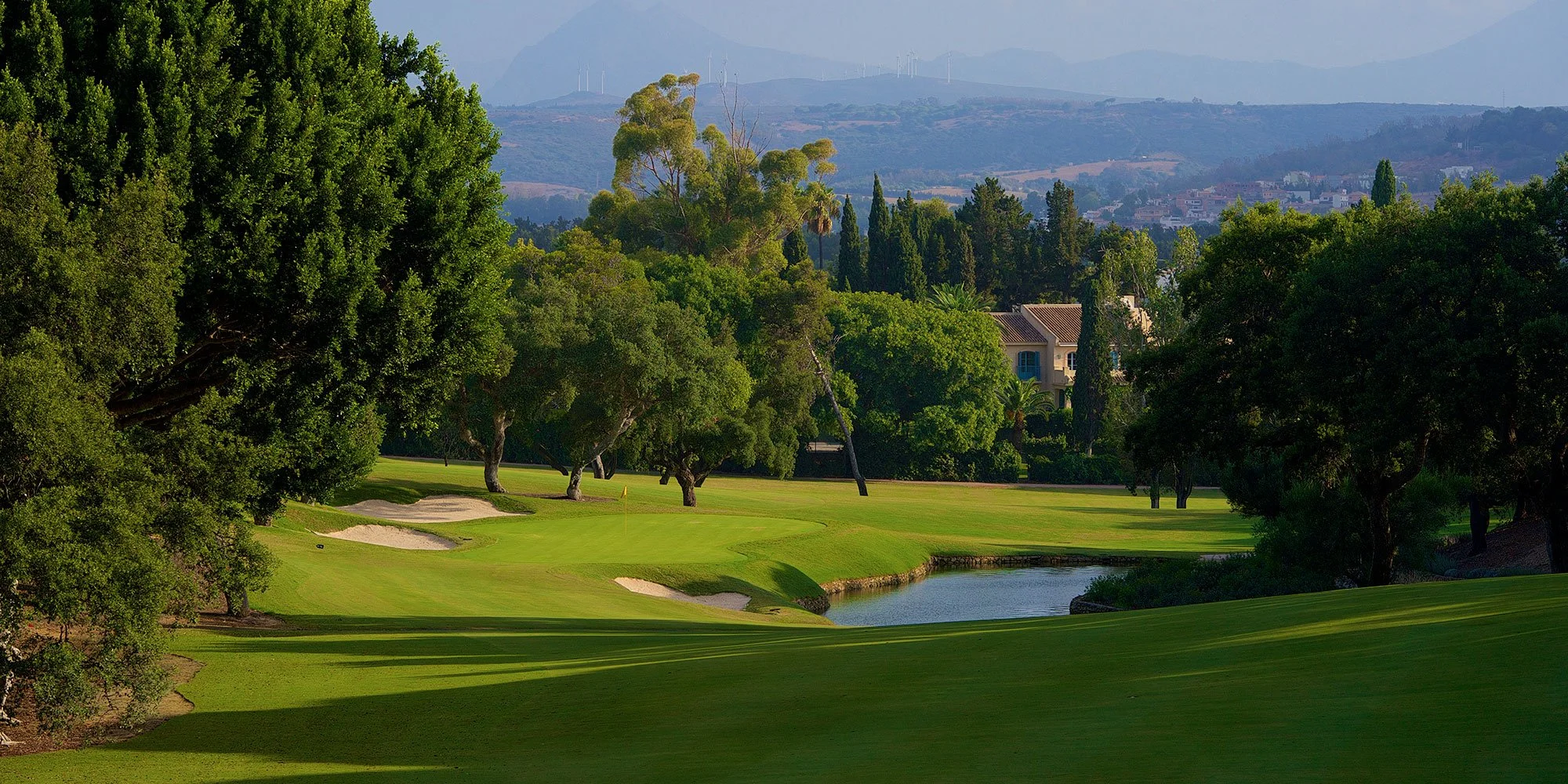 A lush golf course with green fairways, bunkers, water hazards, and trees, with a residential area and distant hills in the background.