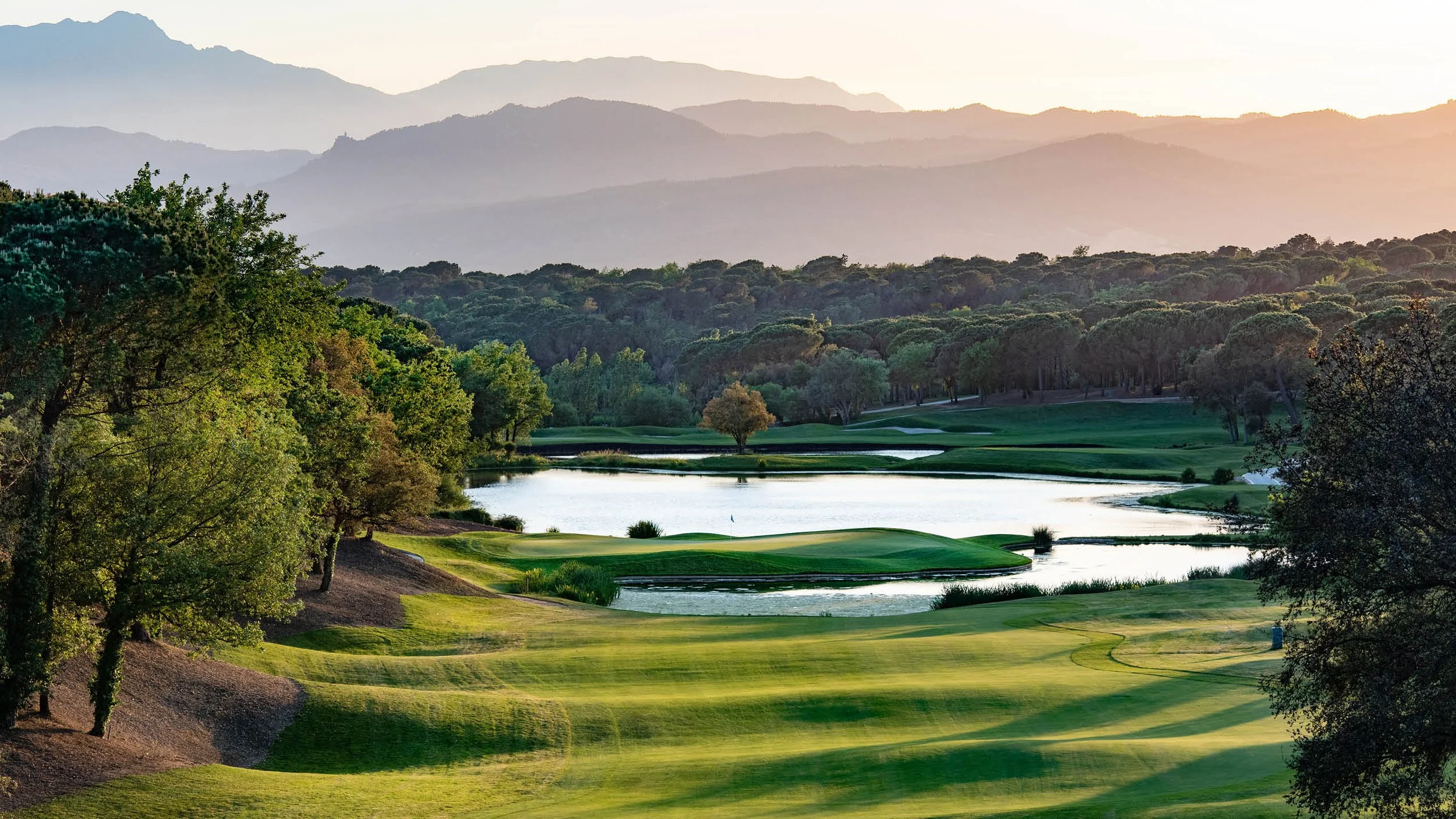 A scenic golf course with water hazards, surrounded by trees and hills, under a hazy sky at sunrise or sunset.