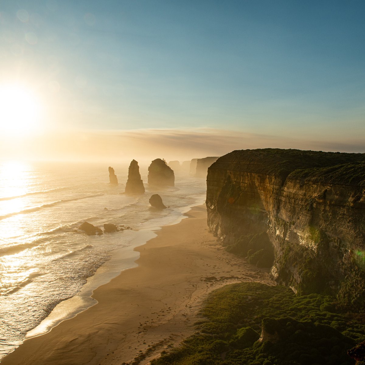 Aerial view of a coastal scene at sunset showing boats anchored in calm water, a wooden pier extending into the water, sandy beach, and a tree-lined shoreline with houses in the background.