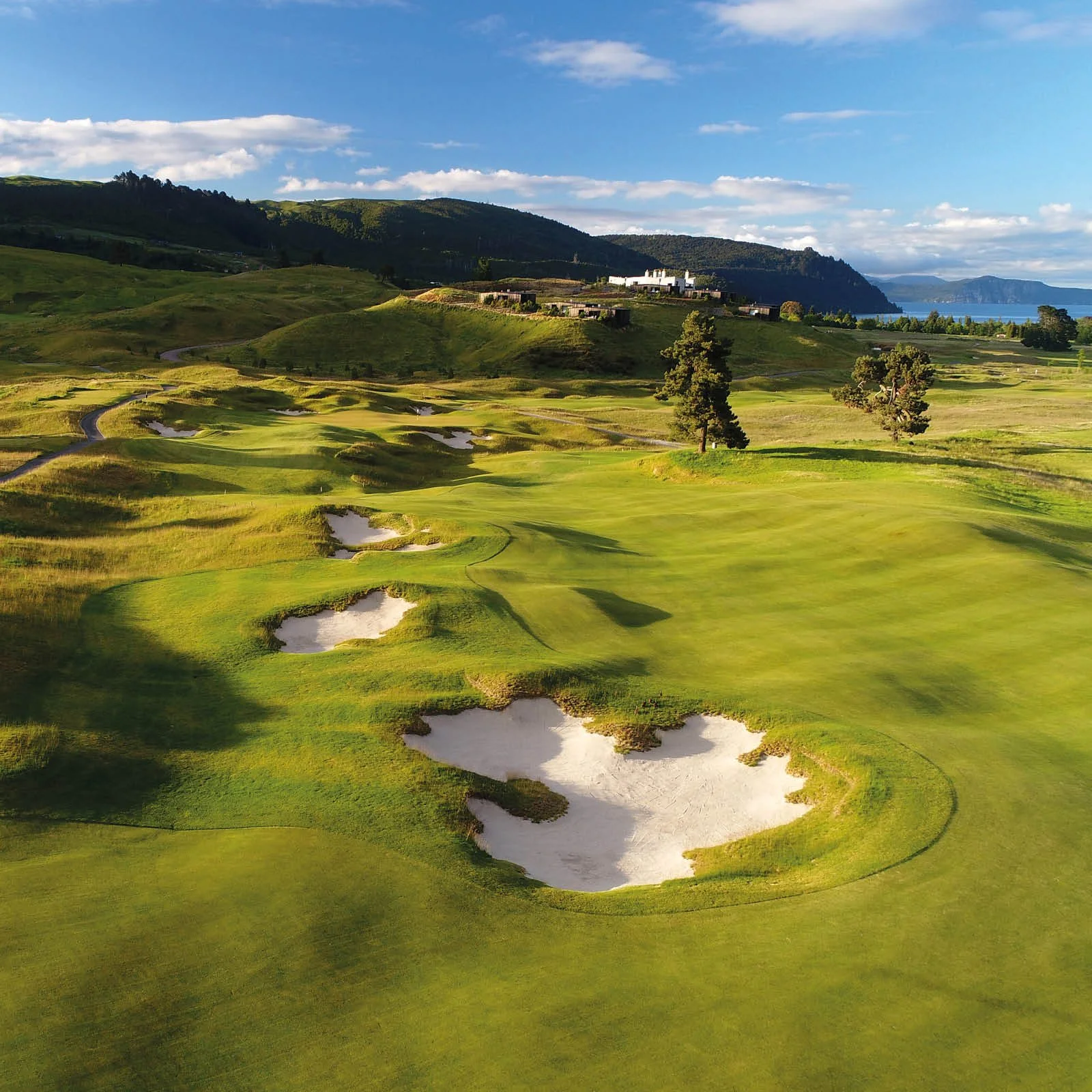 A scenic golf course with sand traps, lush green fairways, and trees, set against rolling hills and a distant coastline under a partly cloudy sky.