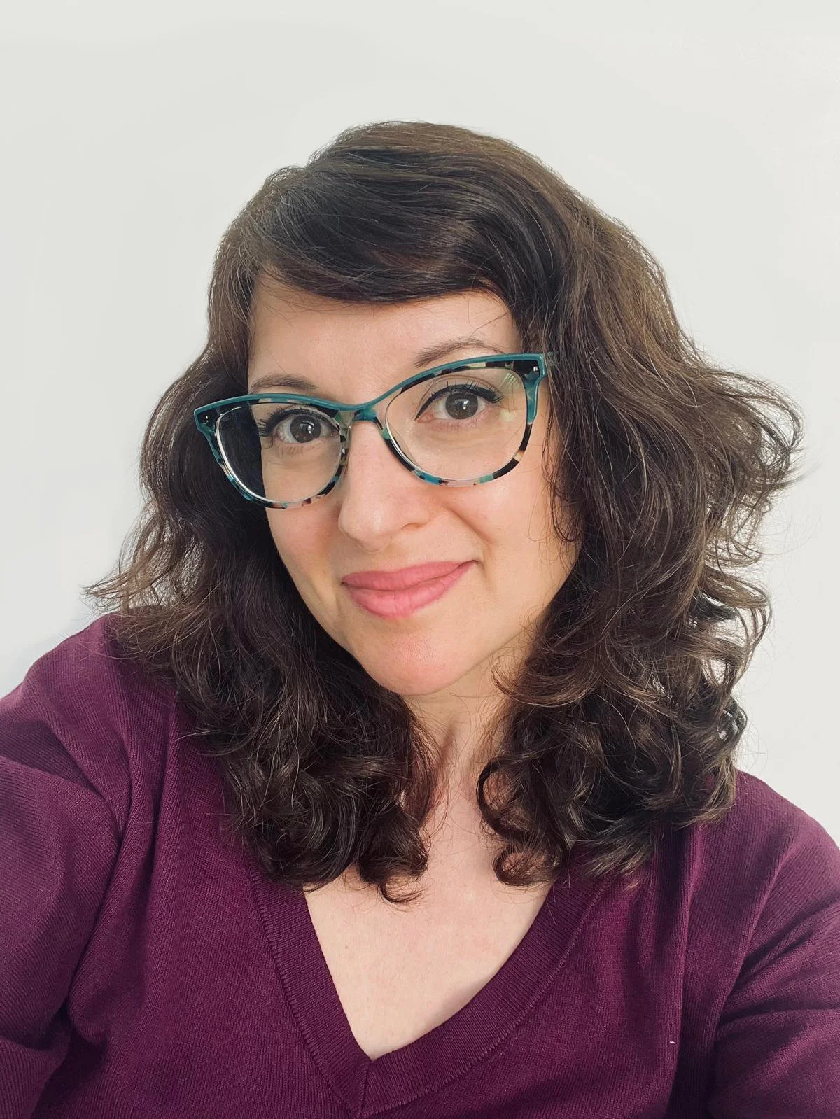 White woman with brown wavy hair and turquoise eyeglasses in a plun v-necked shirt.
