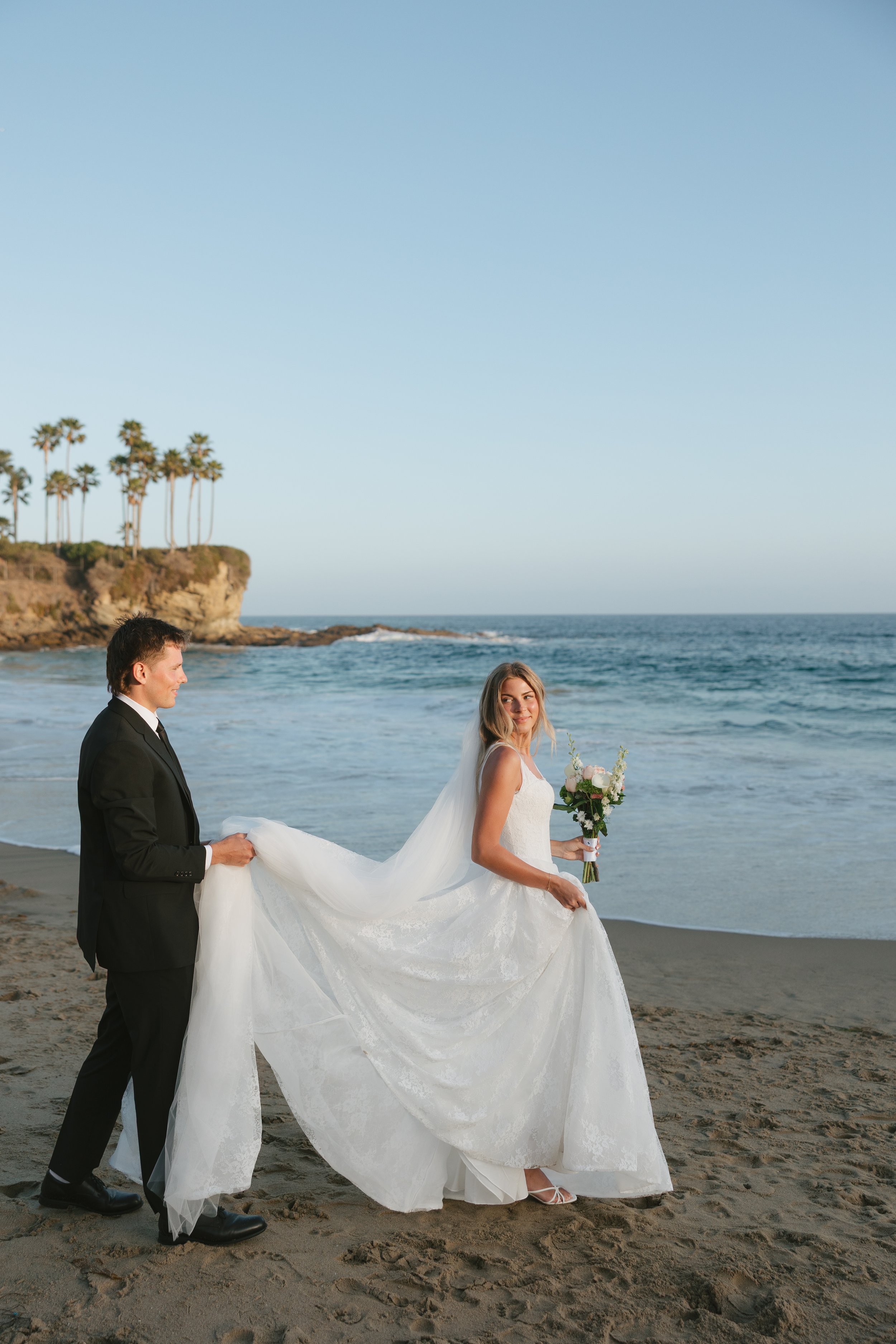 groom helping bride carry her dress on the beach