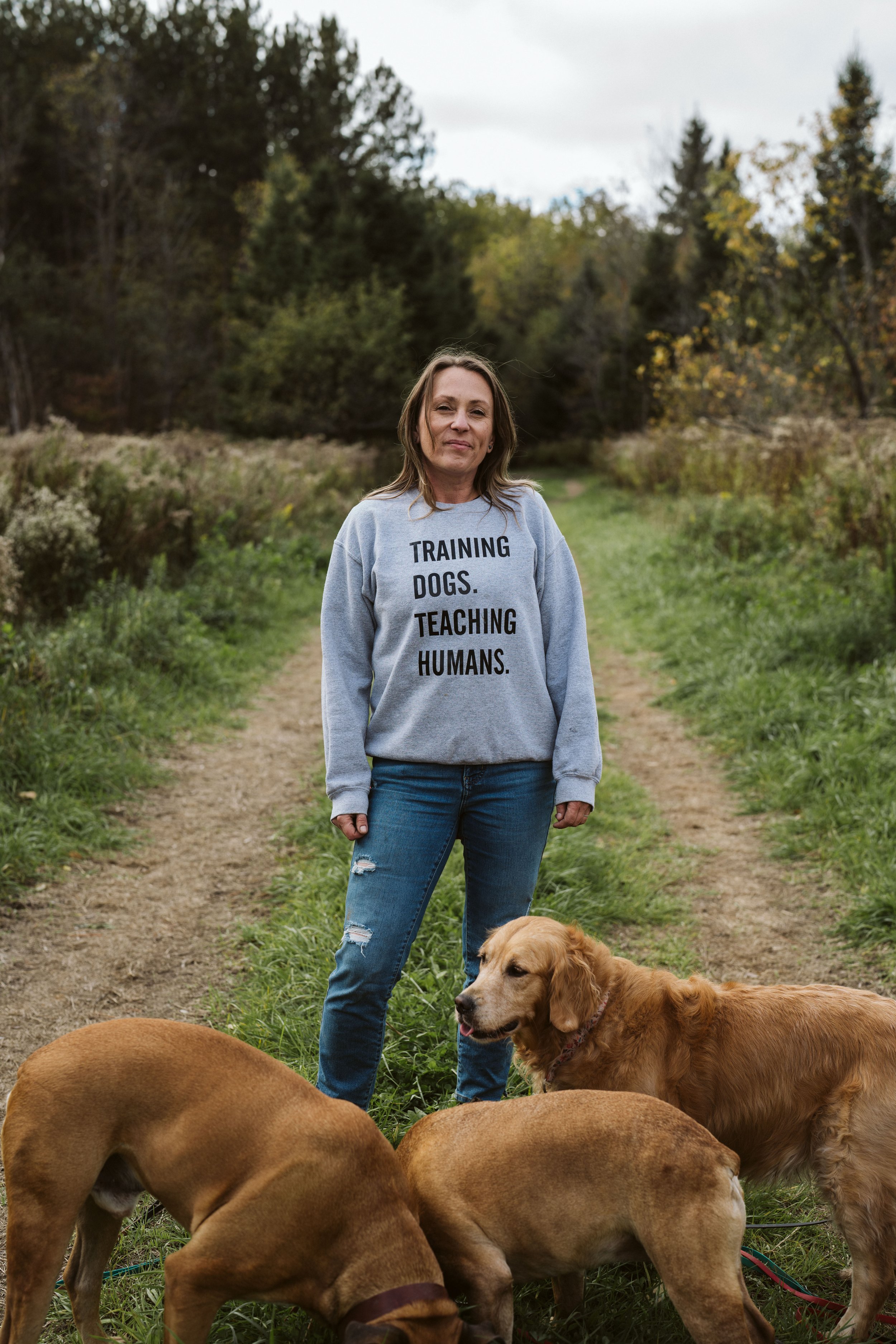Woman standing in field with her tan and red dogs wearing a shirt that says training dogs, teaching humans