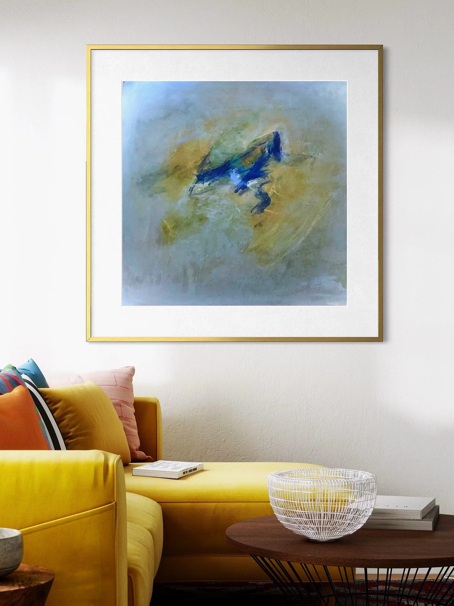 A yellow sofa with colorful pillows in front of a white wall with a large abstract painting in shades of blue, yellow, and green, and a round wooden coffee table with a wire bowl and a few books.