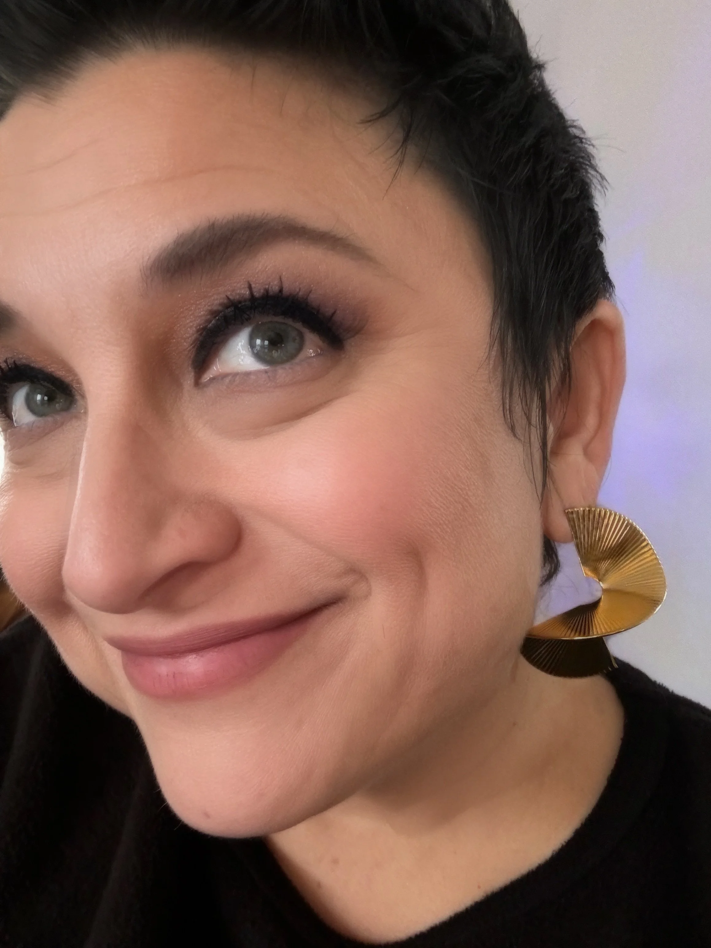 A close-up of a woman with short dark hair, wearing gold earrings, smiling gently, with makeup emphasizing her eyes and cheeks.
