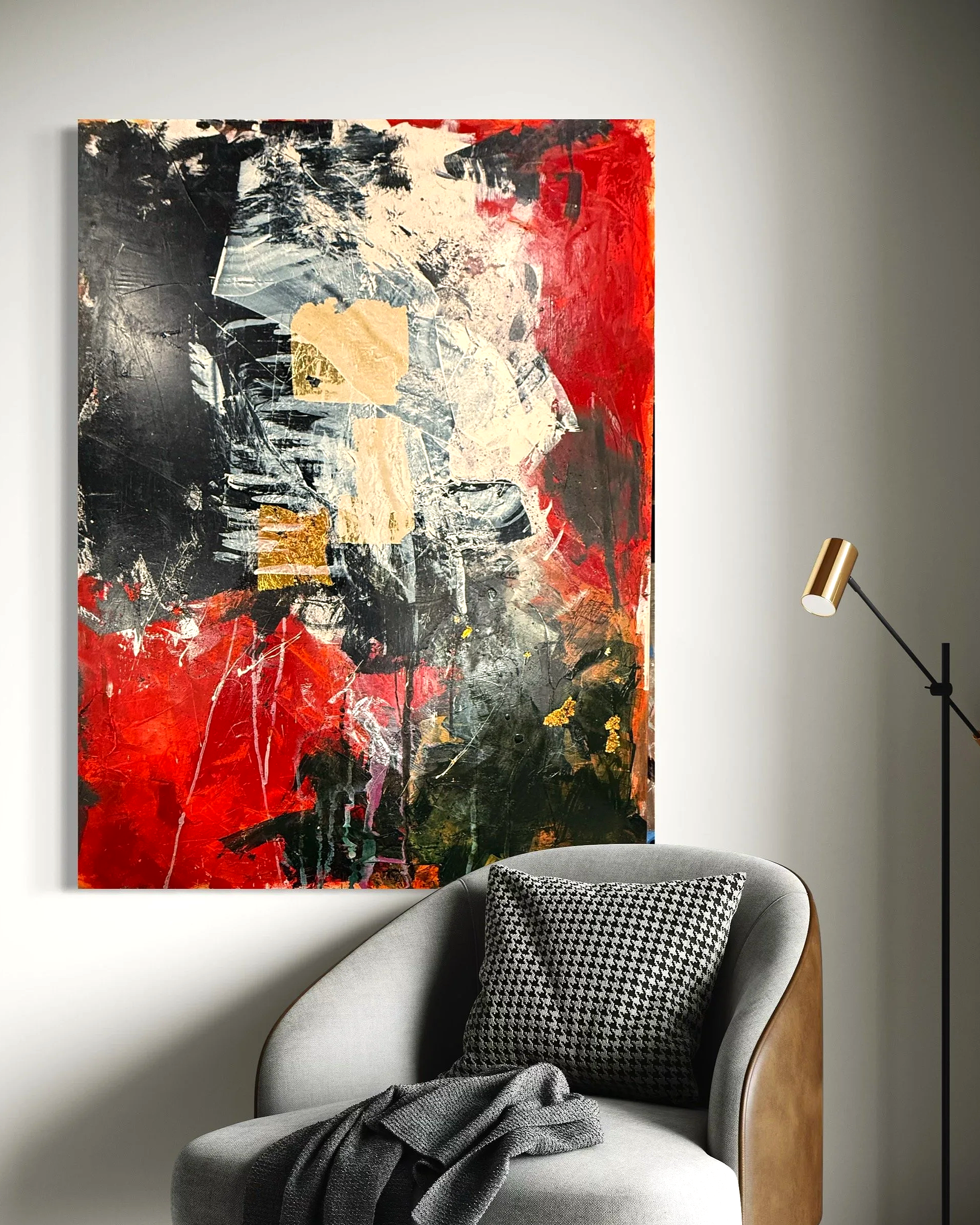 Abstract painting with black, red, beige, and gold colors hanging on a white wall, beside a gray armchair with a black and white pillow and a gray throw, and a tall, black and gold floor lamp.