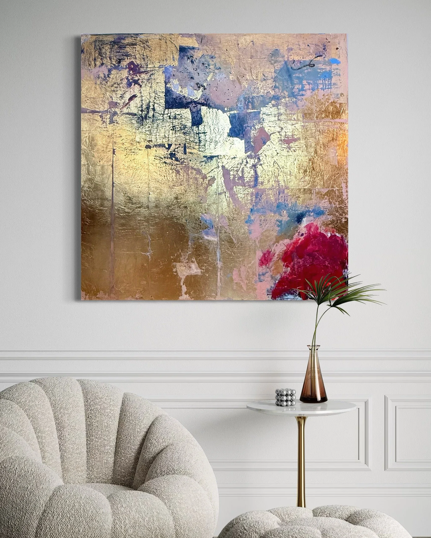 Abstract painting with gold, blue, pink, and red hues hanging on a white wall in a modern living room, with a white textured armchair, a small white table with a glass vase and a decorative silver object.