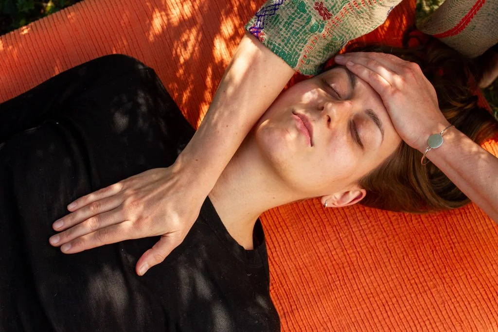 Een vrouw ligt op haar rug op een oranje mat, haar ogen dicht en haar hand op haar voorhoofd, tijdens een ontspanningsmoment of massage.