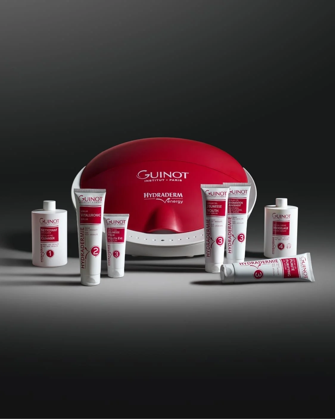 Collection of skincare products from Guinot, arranged in front of a red and white device with a domed shape, on a dark background.