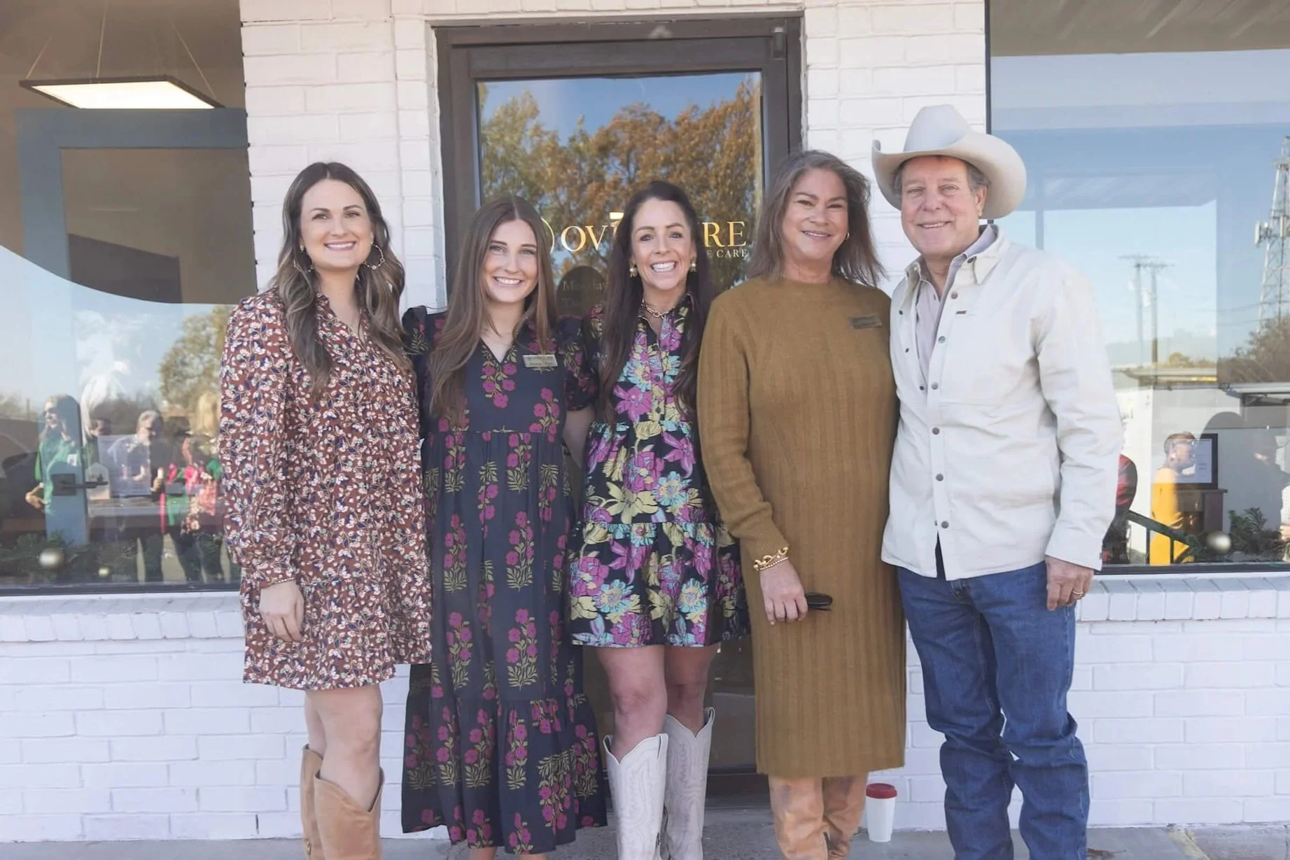 Five people standing in front of a storefront, smiling at the camera. Four women and one man, all dressed in casual to semi-formal clothing. The man is wearing a cowboy hat and a white shirt. The women are wearing floral dresses or skirts, with two of them wearing boots. The background shows a glass door with trees and power lines reflected in it and some people visible through the window.